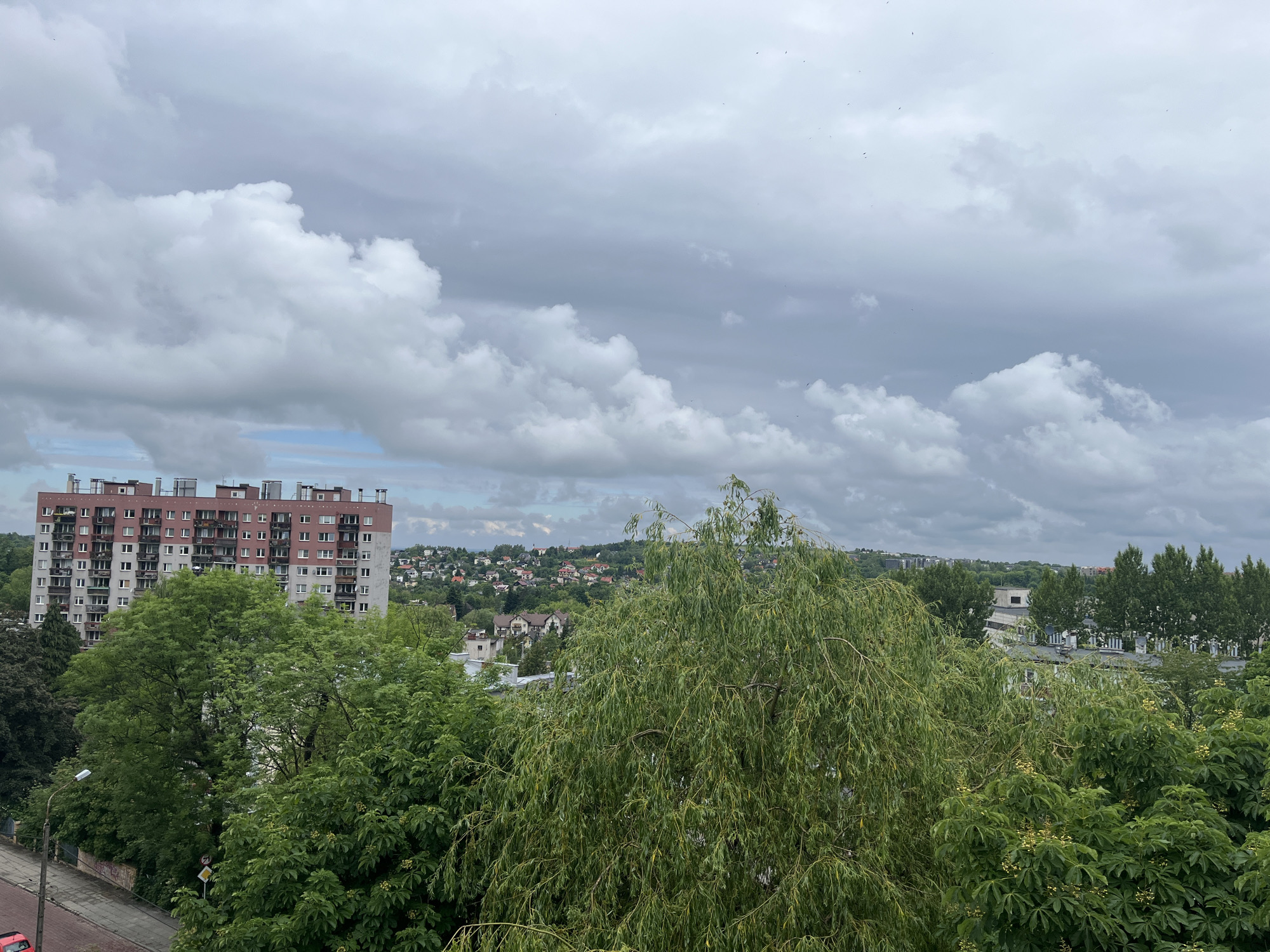 The image captures an elevated view of an urban or suburban landscape under an overcast sky. In the left midground, a tall, multi-story residential building with a pinkish-brown and light gray facade stands prominently. Its roof features numerous vents or chimneys. To the left of this building, a paved street with a sidewalk is partially visible, and a small, red car is parked at the bottom left. A streetlamp and a yellow diamond-shaped traffic sign are also discernible among the trees on the left.
The midground and foreground are dominated by a lush canopy of green trees, including a large weeping willow-like tree on the right. Some trees show signs of light yellow blossoms. Beyond the residential building, in the background, a hillside covered with numerous smaller houses and buildings, characteristic of a town or village, extends into the distance. Further back, more distant buildings and trees are faintly visible against the horizon.
The sky is heavily overcast with thick, scattered cumulus clouds, ranging from bright white to darker gray, suggesting a cloudy day, possibly before rain. Numerous tiny dark specks, likely birds or insects, are scattered across the sky. The scene is static, with no discernible human activity. It is daytime, characterized by diffused light. There is no visible text in the image.