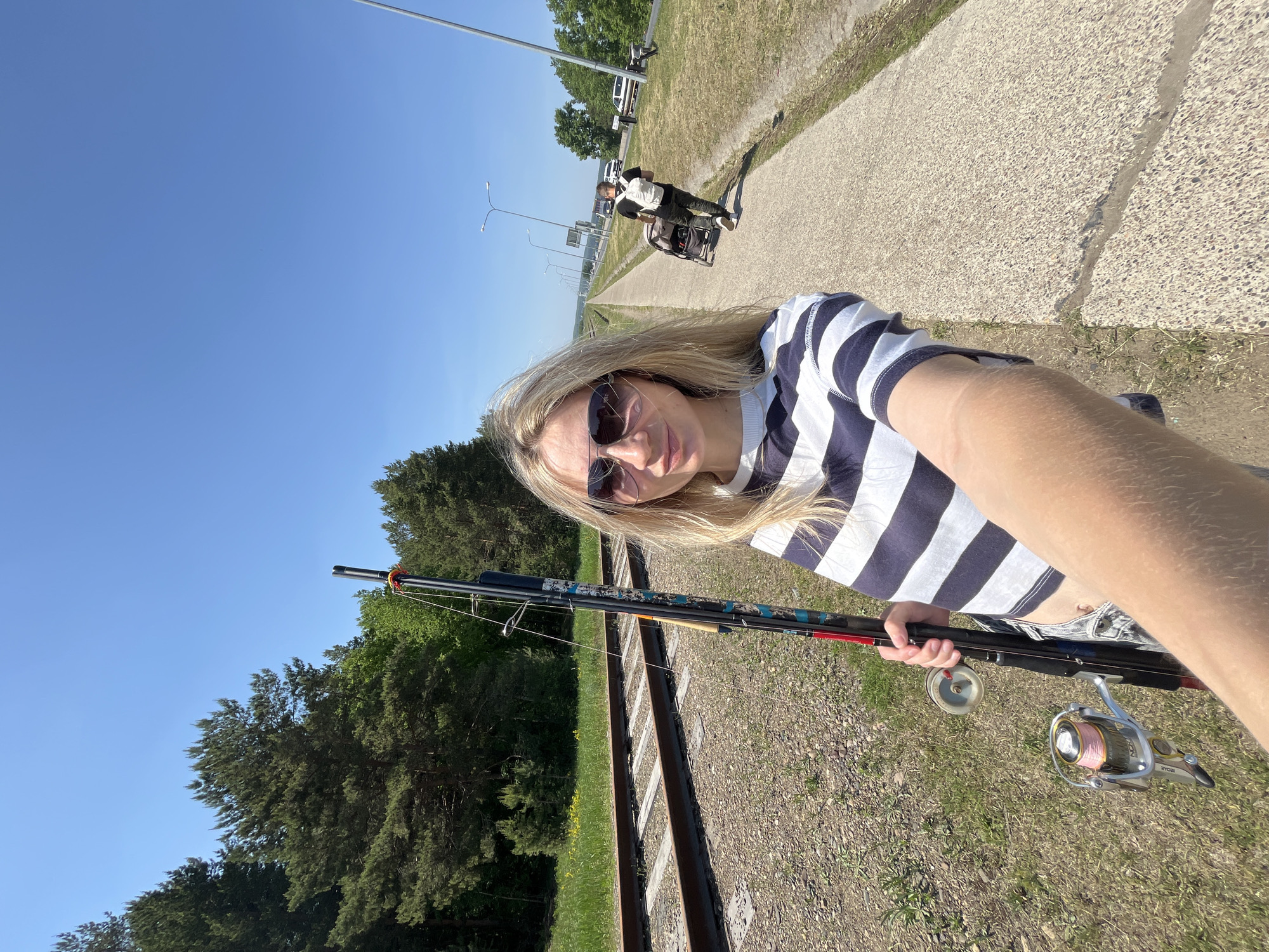 A medium-range outdoor shot shows a blonde woman standing next to railroad tracks, holding fishing poles, with a clear blue sky and a paved path with people in the background.
The woman is fair-skinned, wearing sunglasses, and a horizontally striped navy and white shirt. She is holding the fishing poles in her left hand and appears to be taking a selfie with her right. Two fishing reels are visible.
The railroad tracks run diagonally across the lower portion of the image, with gravel and grass between and around the tracks. Green trees line the left side of the image.
In the background, a paved path runs parallel to a road. A man is walking down the path, pushing a baby stroller. A vehicle is visible on the road. A streetlight is visible on the side of the road, and other streetlights can be seen in the distance.
The city is Chaikovsky, Russia.