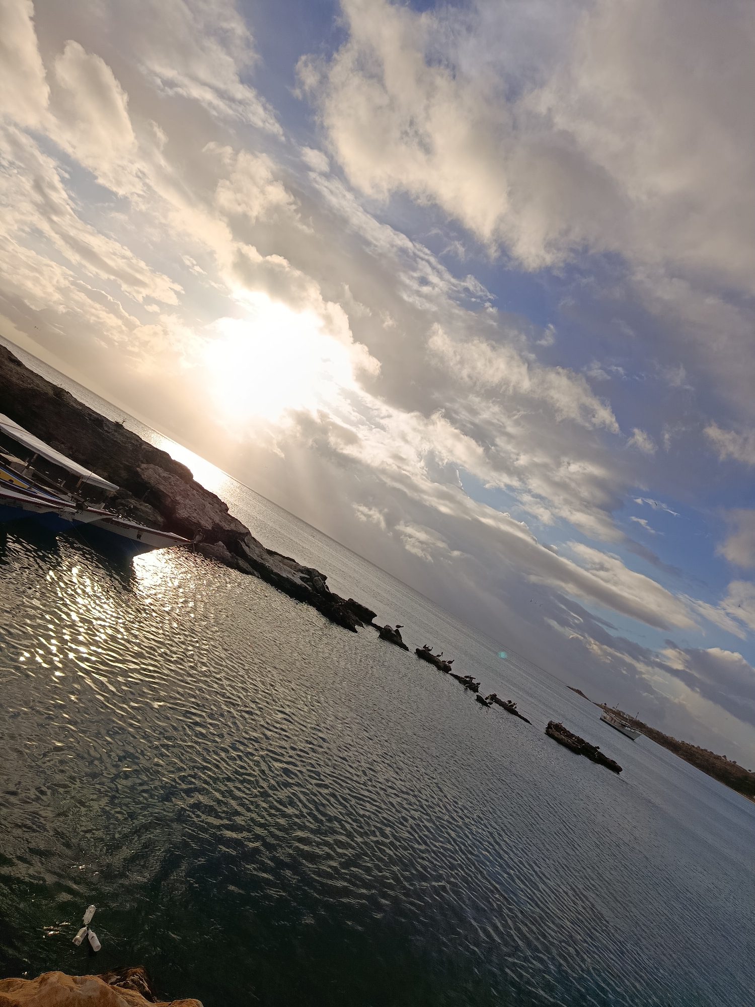 The image depicts a coastal scene in Juan Griego, Venezuela, featuring a body of water with boats moored near a rocky shoreline. The sun is low in the sky, casting a warm glow on the water and illuminating the clouds. Several small boats, likely fishing vessels, are docked along the rocky edge. Some rocks are visible in the water, with birds perched on them. In the distance, a larger white boat is sailing on the sea. The sky is a mix of blue and white clouds, with sunlight breaking through. The water surface shows ripples and reflections from the sun. The overall impression is of a serene coastal landscape at either sunrise or sunset.
