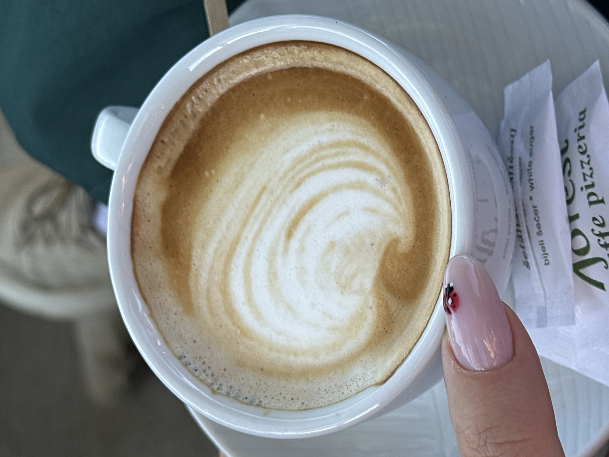 The image shows a close-up, top-down view of a cappuccino in a white ceramic mug, presumably taken at or near the Aorest caffe pizzeria in Šušanj, Montenegro, suggested by the branded sugar packets visible. The cappuccino has a detailed latte art swirl on top. A portion of a person's thumb and fingernail is visible on the right side of the mug, featuring nail art with a red and black ladybug design. In the background, there are partial views of a table and a person's clothing, suggesting an outdoor or café setting.