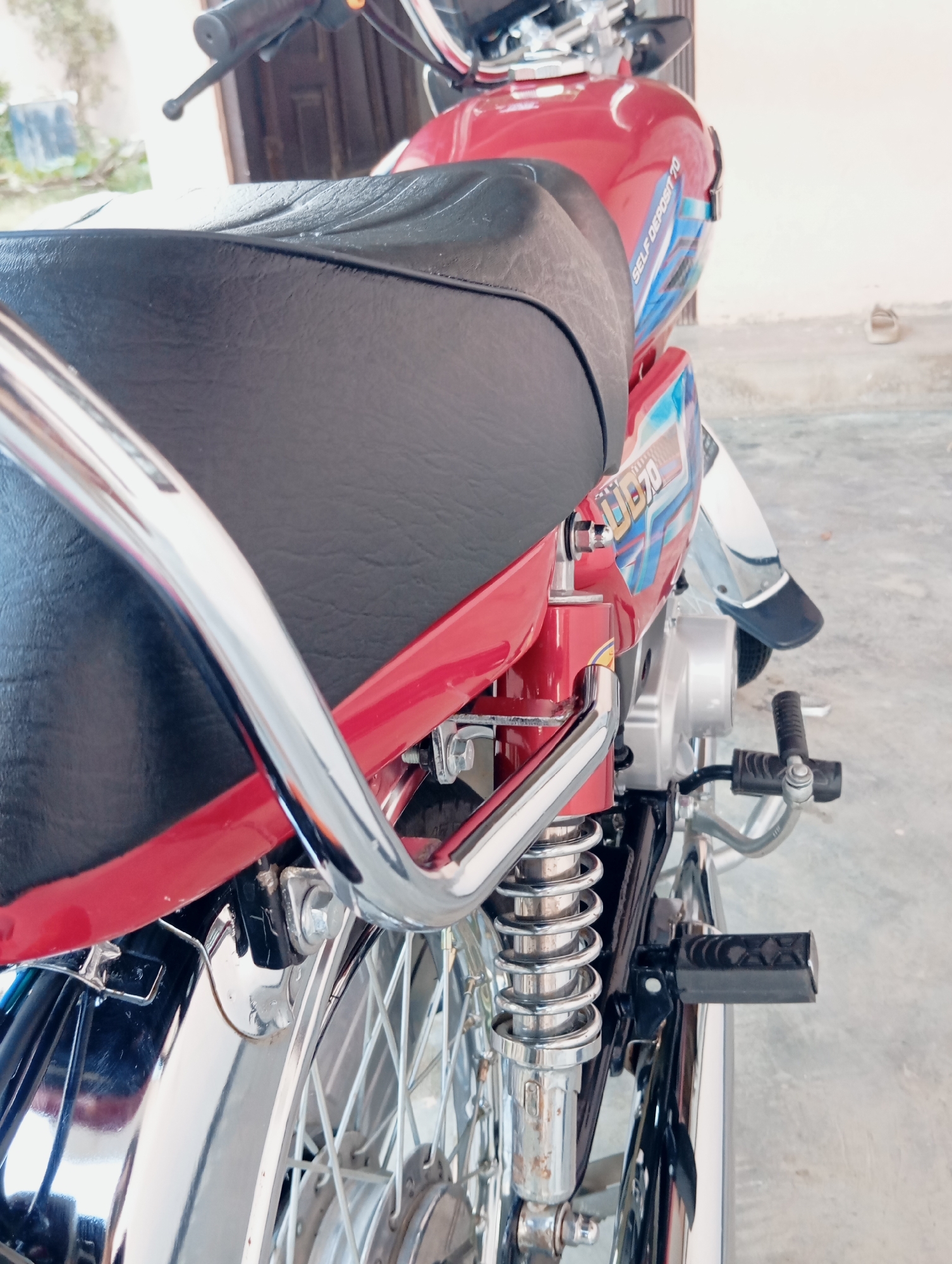 The image is a close-up, side-view, eye-level shot of the rear portion of a red motorcycle. The motorcycle has a black seat and chrome detailing. The brand name "UB 70" and "SELF DEPOSIT 70" are visible on the fuel tank. The background is blurred but appears to be an outdoor setting, possibly a driveway or patio, with a beige wall and a suggestion of foliage and a door in the distance. A pair of beige shoes are visible on the ground to the right of the motorcycle. The motorcycle is stationary and no individuals are present in the frame. The lighting is bright, suggesting daylight.