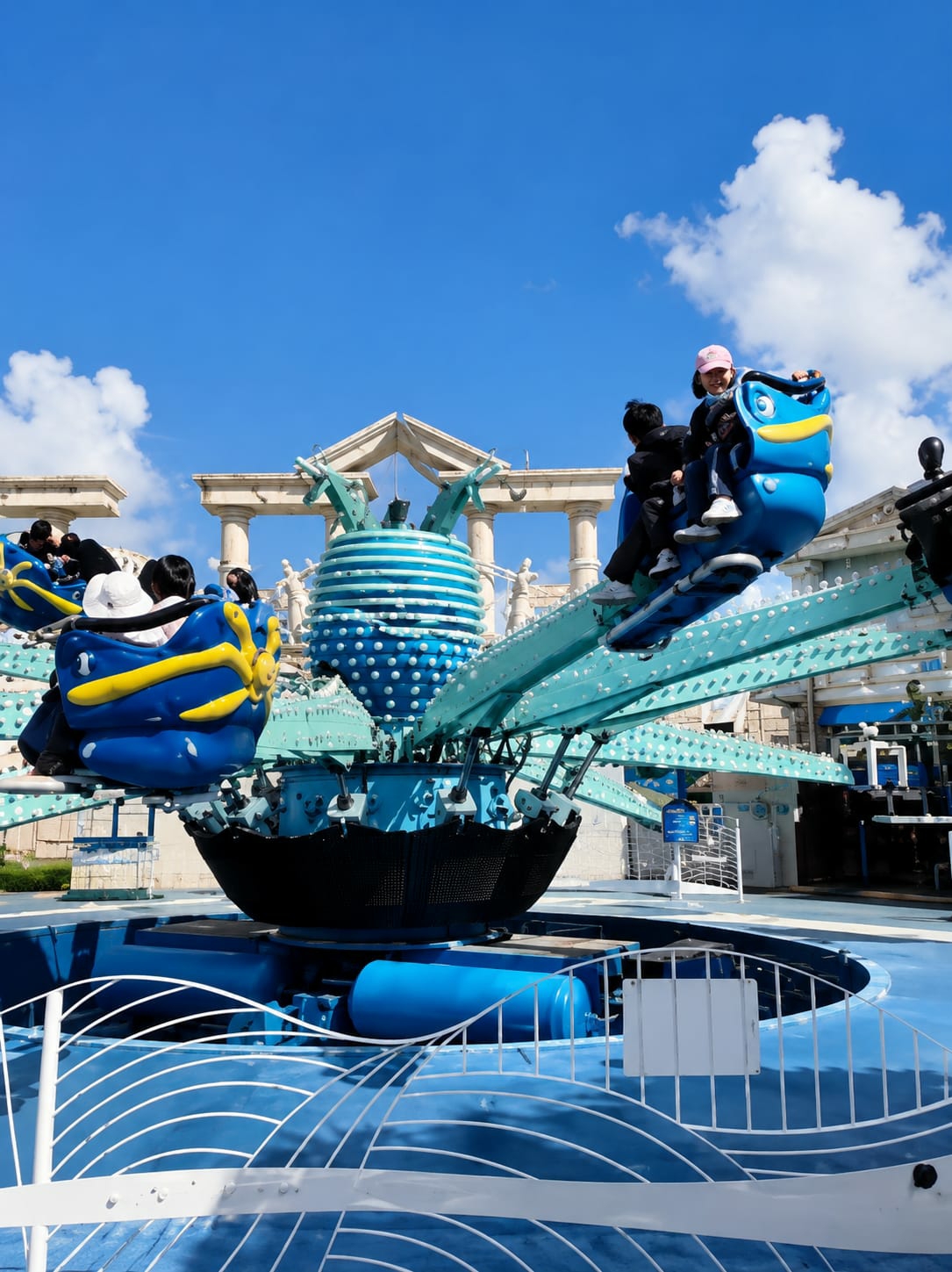 In Berastagi, Indonesia, a brightly colored amusement park ride, possibly a spinning octopus or similar attraction, is in motion under a clear blue sky with scattered white clouds. The ride features several blue gondolas, shaped like cartoonish fish, each carrying passengers. In one gondola, two children are visible: a girl wearing a pink cap and a boy in a dark jacket. The ride itself is predominantly turquoise and blue, with spherical elements and metallic arms extending from a central base. In the background, a white structure with columns, resembling a classical temple facade, suggests the park's architectural theme. The ride's base is surrounded by a blue pool with white wave-like patterns.