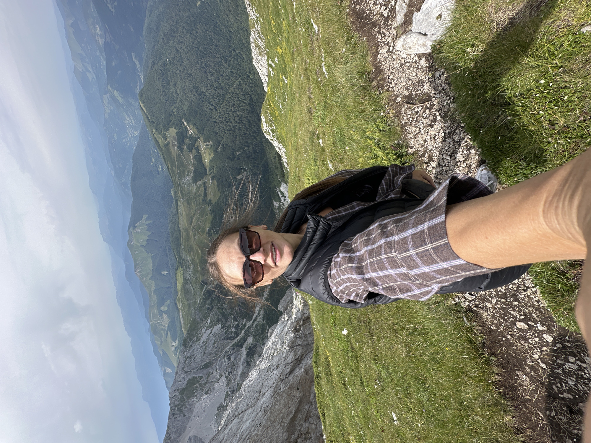 A woman is taking a selfie on a mountain trail in Montenegro. She is wearing sunglasses, a plaid shirt, and a black vest. The background shows a mountainous landscape with green hills and cloudy skies. The trail appears to be rocky and covered in grass.