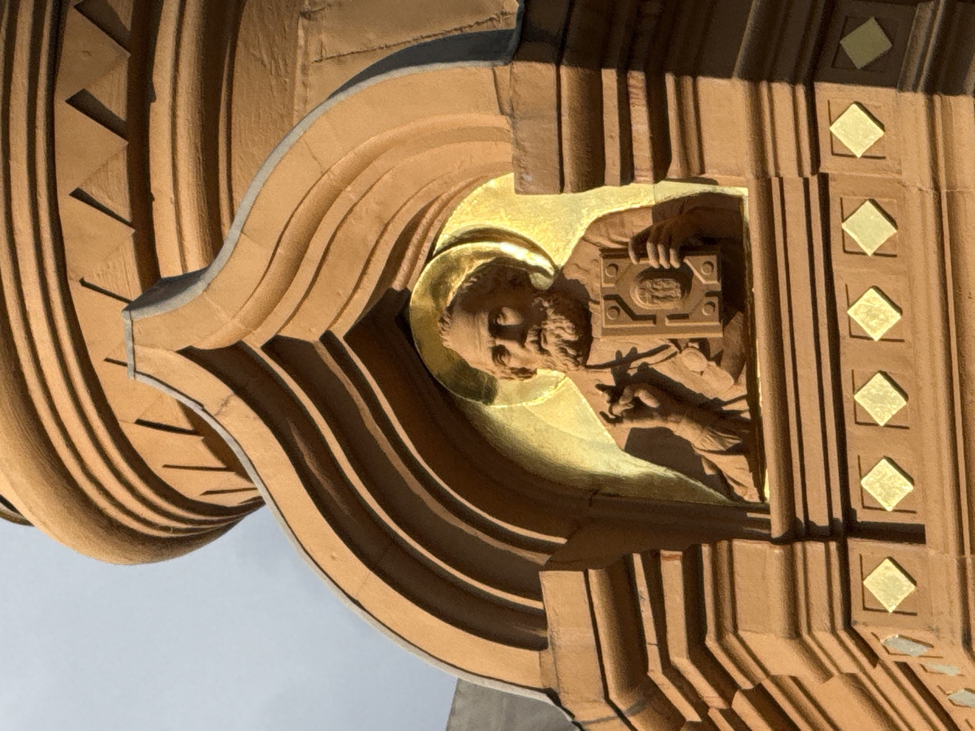 The image shows a close-up of a religious statue carved in relief on the exterior of a building. The statue depicts a bearded man with a halo around his head, holding a book. He is wearing a robe and his right hand is raised in a gesture of blessing. The statue is set within an arched niche, and the surrounding architecture is elaborately decorated with concentric curves and triangular patterns. The material appears to be reddish-brown stone, possibly sandstone or brick, with gold accents on the halo and in diamond-shaped inlays below the niche. The overall impression is of an ornate, possibly Orthodox Christian, religious building. Based on stylistic elements and architectural context, this could be a detail from a church in Moscow, Russia, such as St. Basil's Cathedral or another historical religious site.