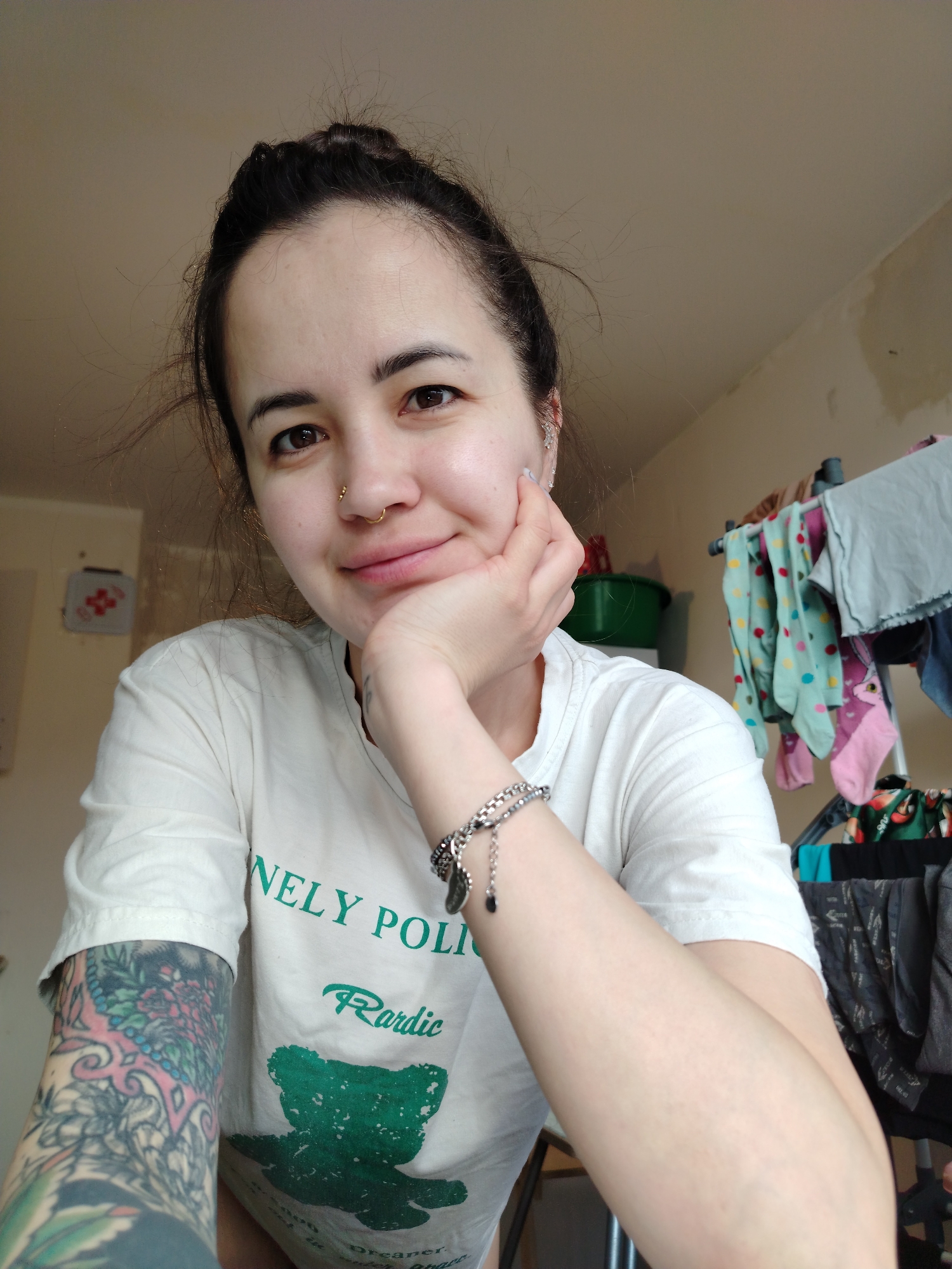 A fair-skinned woman with dark hair pulled into a bun is facing the camera with a slight smile. She has a nose ring and multiple ear piercings. Her left arm is resting on her hand, and a silver bracelet is visible on her wrist. A colorful tattoo covers her left arm. She is wearing a white t-shirt with green text and a graphic. The background includes a partially painted wall, a laundry rack with clothes hanging on it, and a green container on a shelf. The location, city, and country cannot be determined from this image alone.