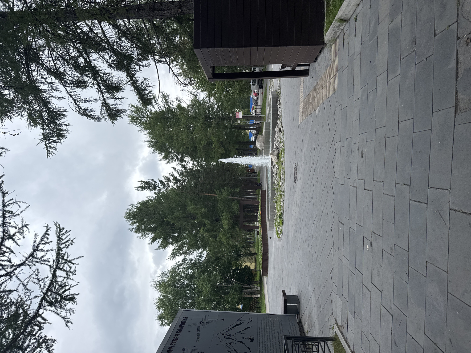 The image depicts an outdoor public space with a fountain as the central feature. The fountain is active, with water spraying upwards from a circular basin surrounded by rocks. The ground is paved with large, grey rectangular tiles, some of which appear slightly uneven. To the left, a dark grey, rectangular informational display board with text and images is visible, accompanied by a grey bench. Several tall, green coniferous trees are scattered throughout the scene, with their branches reaching towards a cloudy sky. On the right side of the image, a dark brown, wooden structure, possibly a shelter or kiosk, stands prominently. Beyond the fountain, a street with parked cars, including a white van and several passenger vehicles, is visible. The overall environment suggests a park or a public square in a city, likely Sosnovka, Russia, given the context. The time of day appears to be daytime, with overcast conditions. There are no people clearly visible in the immediate foreground or interacting with any objects.