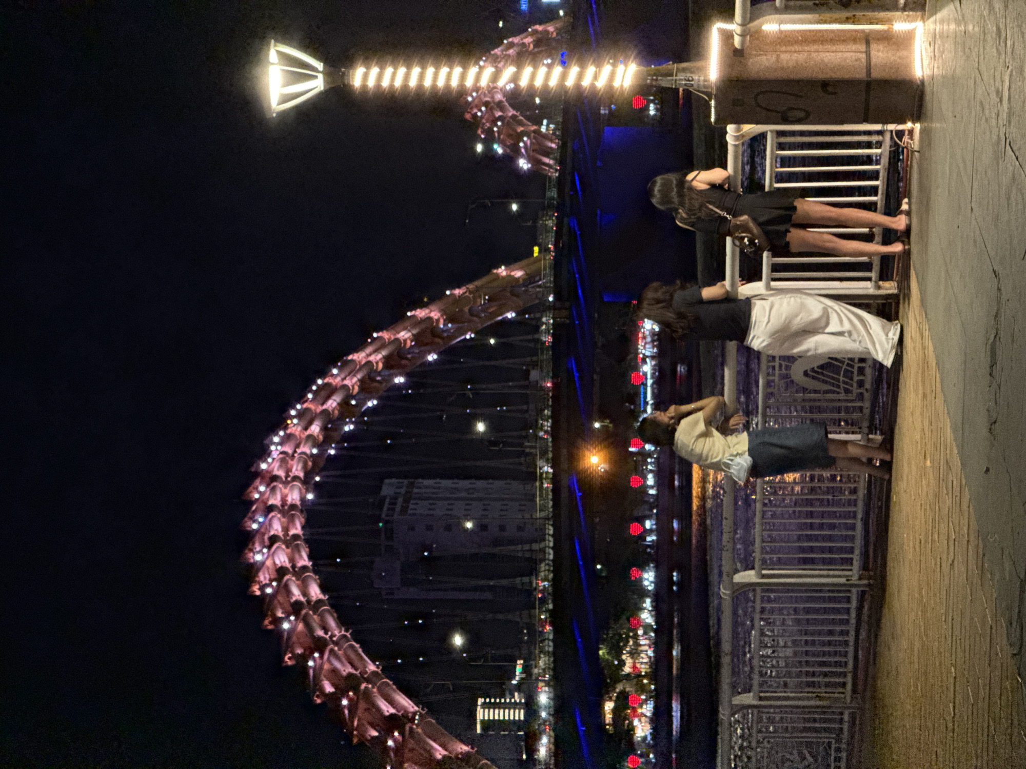 The image shows a nighttime view of the Dragon Bridge in Da Nang, Vietnam. The bridge is illuminated with red and pink lights, resembling a dragon's scales. In the foreground, three women stand by a railing overlooking a body of water. One woman wears a light-colored shirt and a dark skirt, another wears a black top and white pants, and the third wears a black dress. The scene is illuminated by a lamppost on the right side of the frame. The background shows buildings and more lights across the water, reflecting on the surface. The overall atmosphere is one of urban nighttime ambiance.