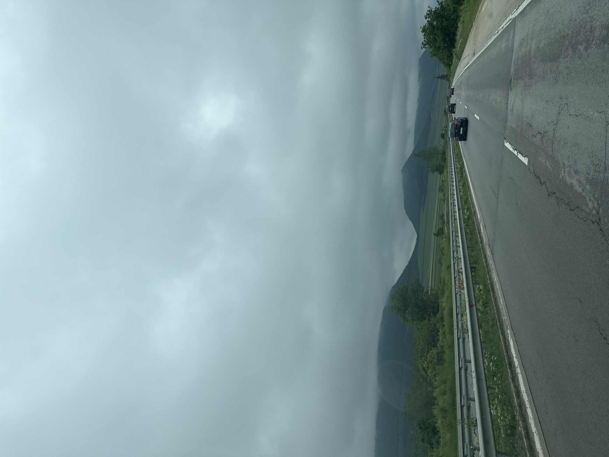 The image depicts a highway near Nevsha, Bulgaria. The road has two lanes with cars traveling in the same direction. A metal guardrail separates the road from the roadside vegetation. Mountains are visible in the background, shrouded in clouds. The sky is overcast.