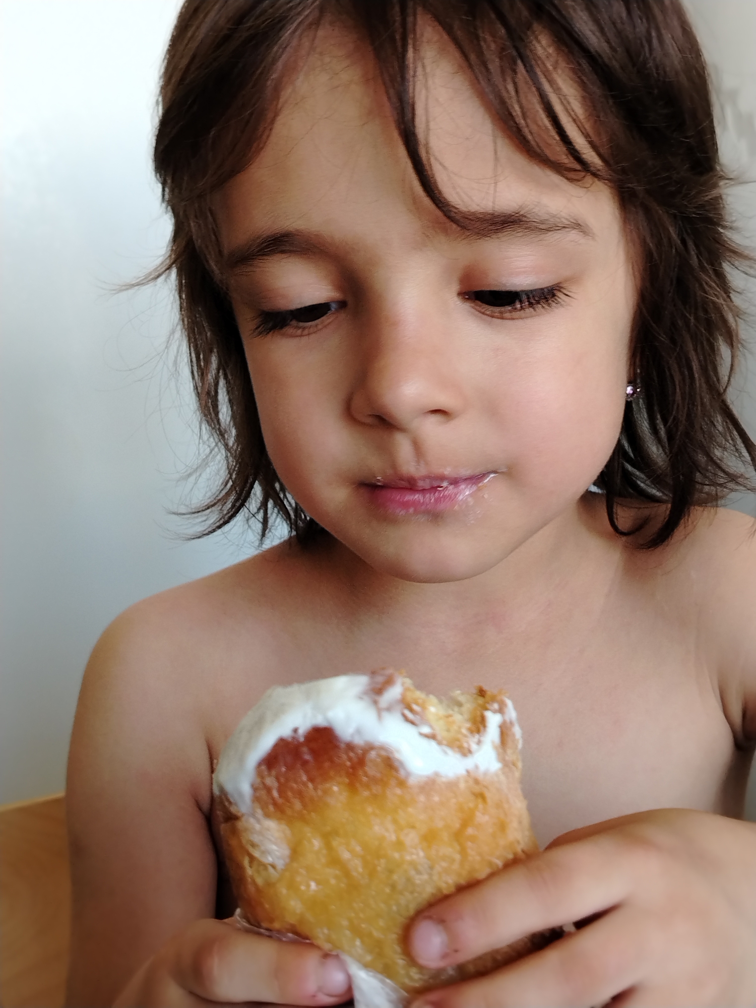 A young child with brown hair and fair skin is holding and looking at a pastry covered in white frosting. The child's lips have traces of the frosting. The child is shirtless and appears to be indoors, with a white wall in the background. The pastry is held in a small hand, and the focus is on the child's expression and the pastry. There is no visible evidence to suggest a location in Noviy, Russia.