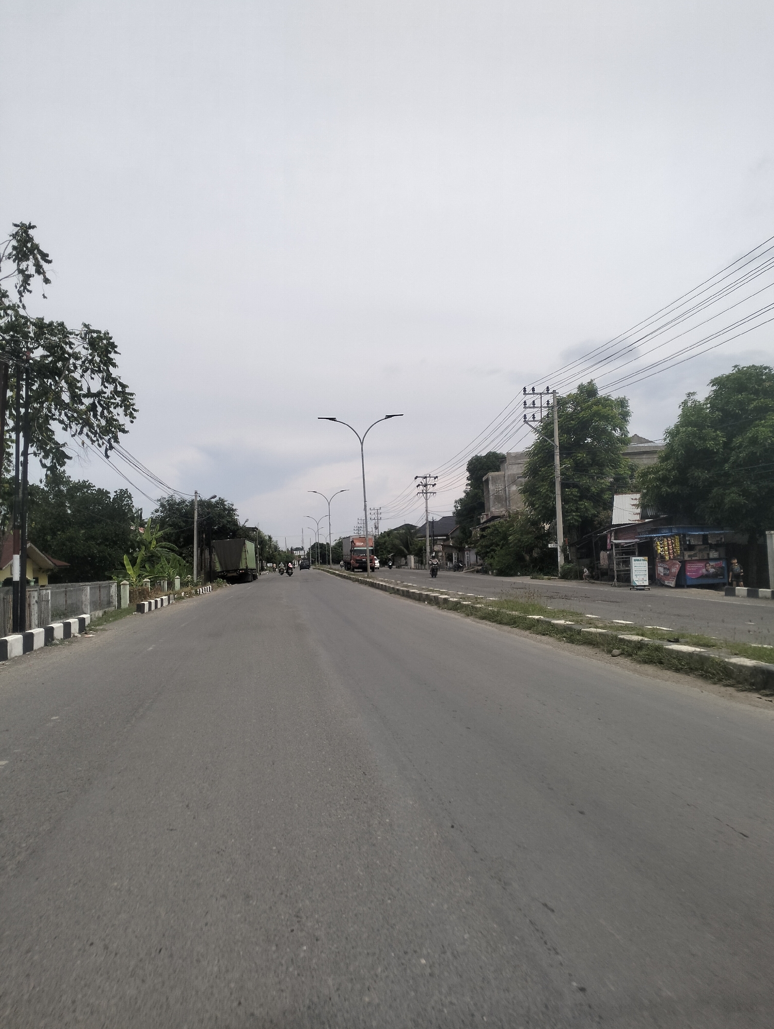 The image depicts a street scene in Lhokseumawe, Indonesia, under an overcast sky. The road is paved asphalt and appears to be a main thoroughfare, stretching into the distance. On the left side of the road, there is a sidewalk with black and white striped borders, followed by a low concrete fence, a house with a brown roof, and dense green trees. On the right side of the road, there are more trees, utility poles with numerous electrical wires, a small shop with goods displayed outside, and a person standing near the shop. Several vehicles are visible on the road, including trucks and motorcycles. One truck is parked on the left side of the road, and another truck and several motorcycles are moving in the distance. The overall atmosphere suggests a moderately busy day in a developing urban or suburban area.