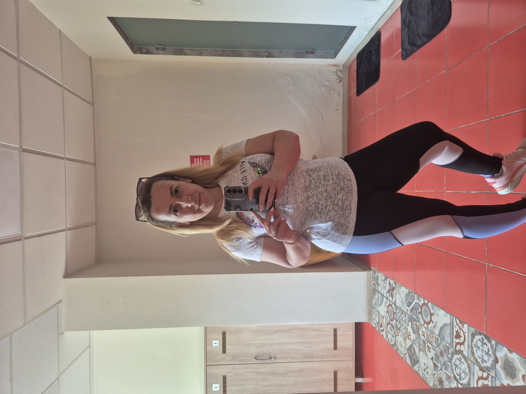 A woman is taking a selfie in a locker room. She is standing in front of a mirror, holding a phone in her right hand. She is wearing a white t-shirt with a graphic print and black leggings with blue and pink accents. She has blonde hair and is wearing sunglasses on top of her head. To her left, there are two wooden lockers. The floor is tiled in red with a decorative pattern of gray and white tiles. To her right, there is a glass-paneled door and two black mats on the floor. The walls are white and appear to have some water damage in places. The ceiling is white and paneled.