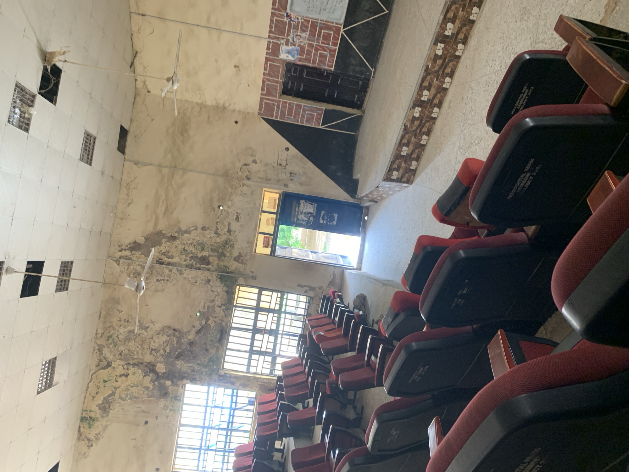 The image depicts the interior of a dilapidated auditorium. The walls show signs of severe decay with peeling paint and water damage. Rows of red and black theater seats are arranged on tiered seating. Two ceiling fans are visible, with one appearing to be in disrepair. The room has large, barred windows that let in natural light. A stage area is visible at the far end of the room, with two doors and a whiteboard. The overall impression is one of neglect and disuse. The location is identified as an auditorium in Jalingo, Nigeria.