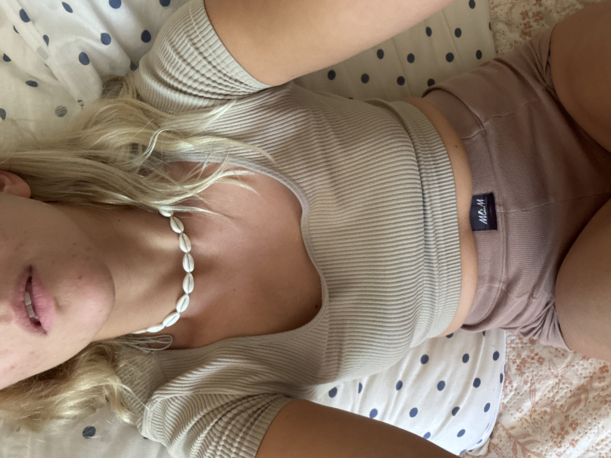 A woman with blond hair is lying on a bed with a white and blue polka dot pillow and a pink floral patterned quilt. She is wearing a ribbed beige top, pink shorts with a black tag labeled "M&M", and a white shell necklace. The environment suggests a bedroom setting. No information is available to place the location in the city of Minsk, Belarus.