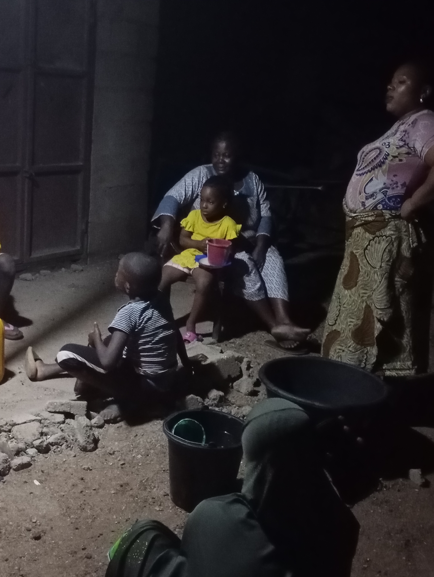 In the city of Shendam, Nigeria, at night, a group of individuals are gathered outdoors, with a building to their left and a dark background suggesting it is nighttime. The ground is composed of dirt and scattered debris, including small rocks and what appear to be broken bricks. To the right, two large dark basins are visible. A black bucket with a green object inside sits in the foreground.
In the center of the scene, an adult woman is seated on a bench, holding a young child on her lap. The child is wearing a bright yellow dress and holding a red bucket. To the left of the seated adult and child, a young boy is sitting on the ground, facing away from the camera. He is wearing a striped shirt and dark pants. To the far right, another adult woman stands, looking upwards and to the right, wearing a patterned dress and a headscarf. In the foreground, partially obscured by darkness, a person wearing a green head covering and a dark garment is seated. The overall atmosphere appears to be a communal gathering in an informal outdoor setting at night.