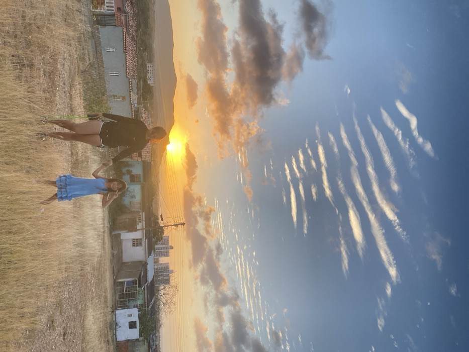 An adult female and a young female child are depicted standing on a dry, grassy hillside overlooking the city of Juan Griego, Venezuela, during sunset. The adult, positioned on the left, wears a dark long-sleeved shirt and dark shorts, holding a bright green object in her left hand. Her right hand clasps the left hand of the child. The child, situated to the right, wears a light blue ruffled dress and holds her right hand in a 'V' gesture. Both individuals face the camera. The immediate foreground consists of dry, straw-colored grass. Below the hillside, the residential area of Juan Griego extends, featuring numerous single-story houses with varied colors and roof types, including terracotta tiles. An electrical pole with multiple wires is visible above these structures. In the background, hills and mountains are silhouetted against a prominent, bright orange-yellow sun setting on the horizon. The sky exhibits a gradient from warm orange and yellow near the horizon to light blue overhead, interspersed with scattered dark clouds and a distinctive pattern of parallel, wave-like white clouds in the upper right. Taller, multi-story buildings are visible in the distant urban landscape.