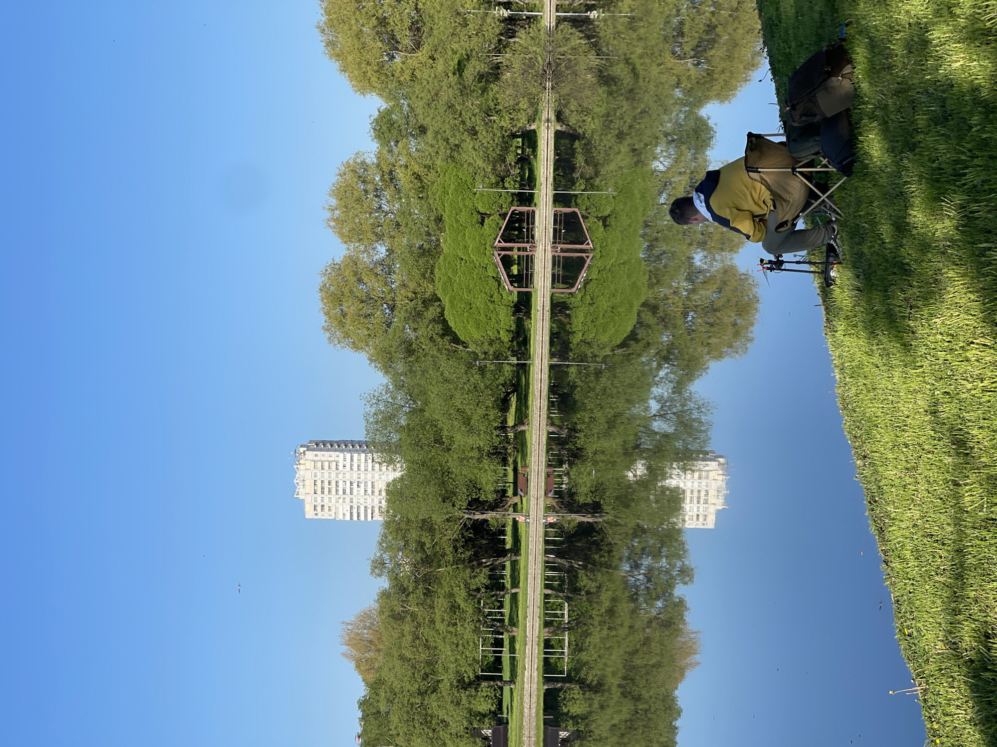 The image is a perspective trick, rotated 90 degrees to the left, making the sky appear as water.
The scene depicts a park or green area, likely next to a body of water, given the reflected image. A man is sitting on a folding chair on the grass, with fishing equipment set up, suggesting he is fishing. The man wears a yellow and blue jacket.
Across the "water", trees line what is presumably a path. There's a building visible in the distance.
The building resembles the Kotelnicheskaya Embankment Building, one of the "Seven Sisters" skyscrapers built during the Stalinist era in Moscow, Russia. Given the architectural style and location, this could likely place the scene in or around the Moscow region.