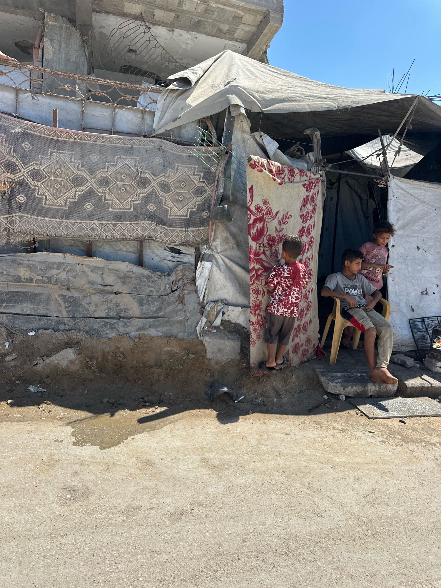 In Gaza City, Palestinian Territories, a group of three children stand outside a makeshift dwelling constructed from tarpaulins and salvaged materials, attached to the remains of a damaged building. The building itself shows signs of destruction, with exposed rebar and cracked concrete. A large patterned rug hangs as a curtain over the entrance. The ground in front of the dwelling is unpaved and shows a puddle of water, suggesting recent rainfall or leakage. One boy, wearing a grey t-shirt and khaki pants with red accents, sits on a yellow plastic chair, looking downwards. Another boy, in a red and white patterned shirt and brown shorts, stands to the left, facing away from the camera. A younger girl, wearing a pink shirt, stands slightly behind the seated boy, looking towards the left. The overall scene depicts a precarious living situation amidst a backdrop of urban decay and conflict.