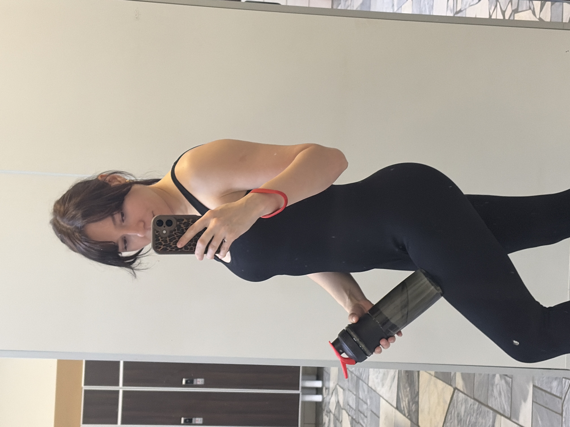 A young woman, with dark brown hair and bangs, stands in what appears to be a gym locker room, taking a mirror selfie. She is wearing a black sleeveless athletic top and black leggings. She is holding a black water bottle with a red lid in her left hand and her phone, with a leopard-print case, in her right hand. A red bracelet is visible on her right wrist. In the background, to the left, are several dark brown gym lockers with silver handles and white bases. The floor has a checkered pattern of light and dark gray tiles. The wall behind her is a light cream color. The overall impression is a candid shot taken in a public fitness facility.
