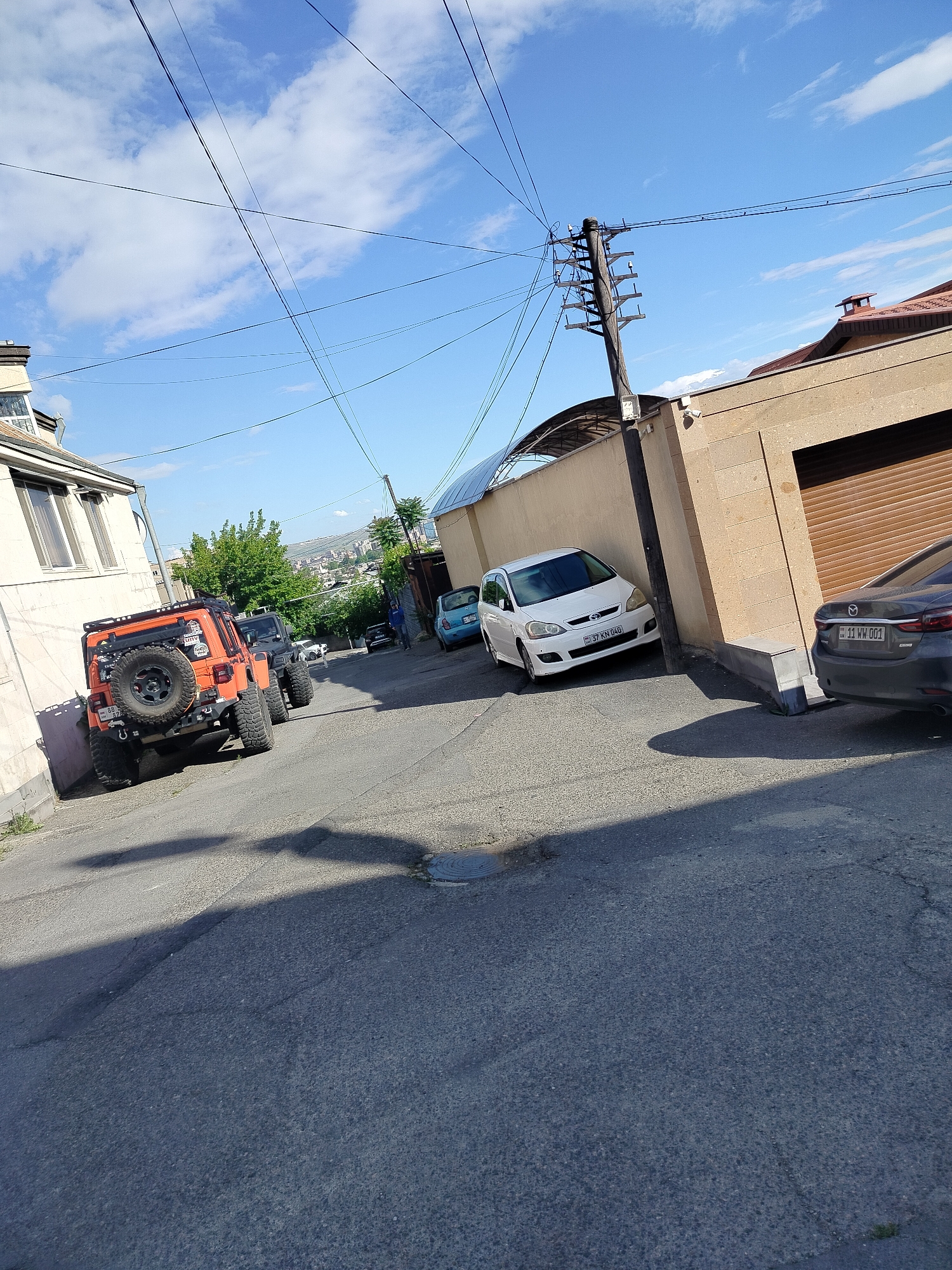 This image captures a street scene in Yerevan, Armenia. The street slopes upwards, with an orange Jeep Wrangler parked on the left side, its rear tire prominently displayed. Behind it, a dark-colored Jeep-like vehicle is partially visible. A white sedan, bearing the license plate "37 KN 040", is parked on the right, adjacent to a tan-colored wall that encloses a building with a brown garage door. A gray Mazda sedan is parked next to the garage door, its license plate reading "11 WW 001". In the distance, other parked cars and a pedestrian can be seen. Overhead, utility poles and crisscrossing wires dominate the sky, which is partly cloudy. The architecture in the background indicates an urban environment.
