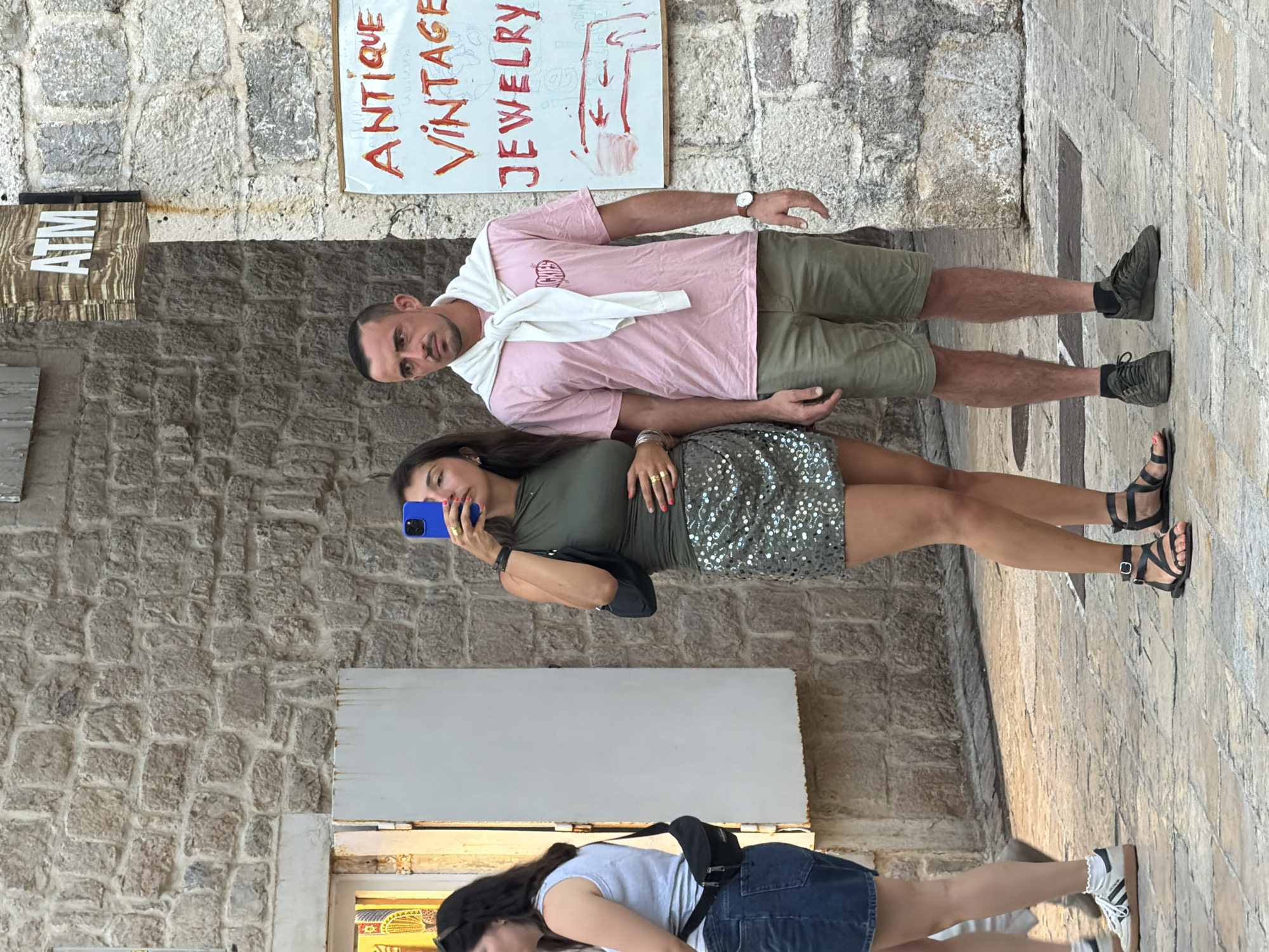 In this vertical, full-body shot, two women and a man are captured outdoors against a stone wall. The man, in the foreground, wears a pink t-shirt and olive-green shorts, with a white sweater draped over his shoulders. He is looking towards the camera. Next to him, a woman in a green top and a sequined skirt is taking a selfie with her blue-framed phone. She is wearing rings on her fingers. Further down and partially visible, another woman in a light blue t-shirt and denim skirt is also captured.
Behind the man, a sign reads "ANTIQUE VINTAGE JEWELRY" in bold lettering. To the left of this sign, another sign with the letters "ATM" is visible. The scene appears to be a street in Budva, Montenegro, with stone walls and paving stones indicating an old town setting. The lighting suggests it is daytime, with diffused sunlight. The overall mood is casual and relaxed, with the subjects engaged in capturing memories of their outing.