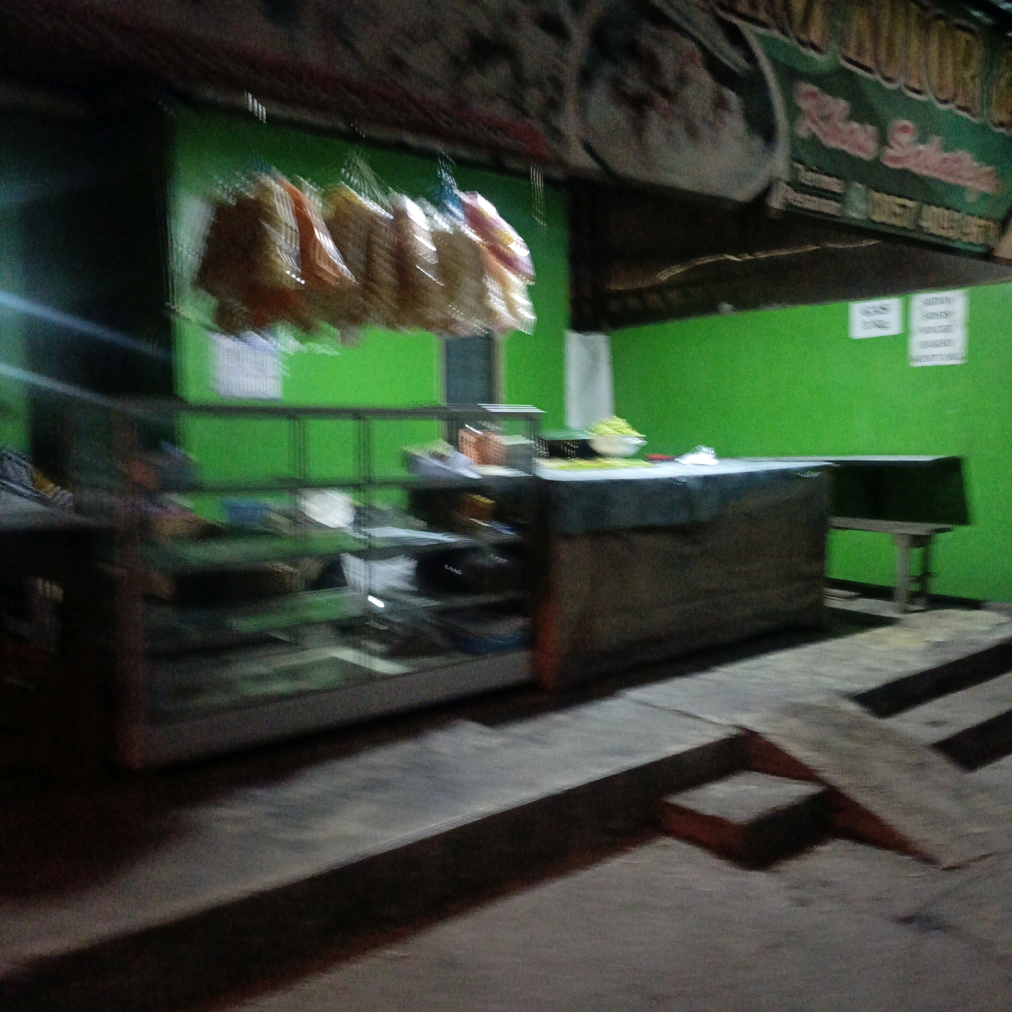 The image depicts a street food stall at night, likely in Salatiga, Indonesia, given the sign's text in Indonesian. The stall features a vibrant green wall as a backdrop. Several packages of snacks, appearing to be crackers or chips, hang from a string in front of the stall. In the center, a display case exhibits various items, though they are blurry. To the right, a counter with a wooden front holds some food items, including a bowl of what appears to be chopped vegetables. A small wooden stool is placed beside the counter. The foreground shows concrete steps leading up to the stall, with some of these steps casting distinct shadows. The overall lighting suggests it is nighttime, with the stall likely illuminated by artificial light. The blurriness of the image indicates motion or a long exposure.