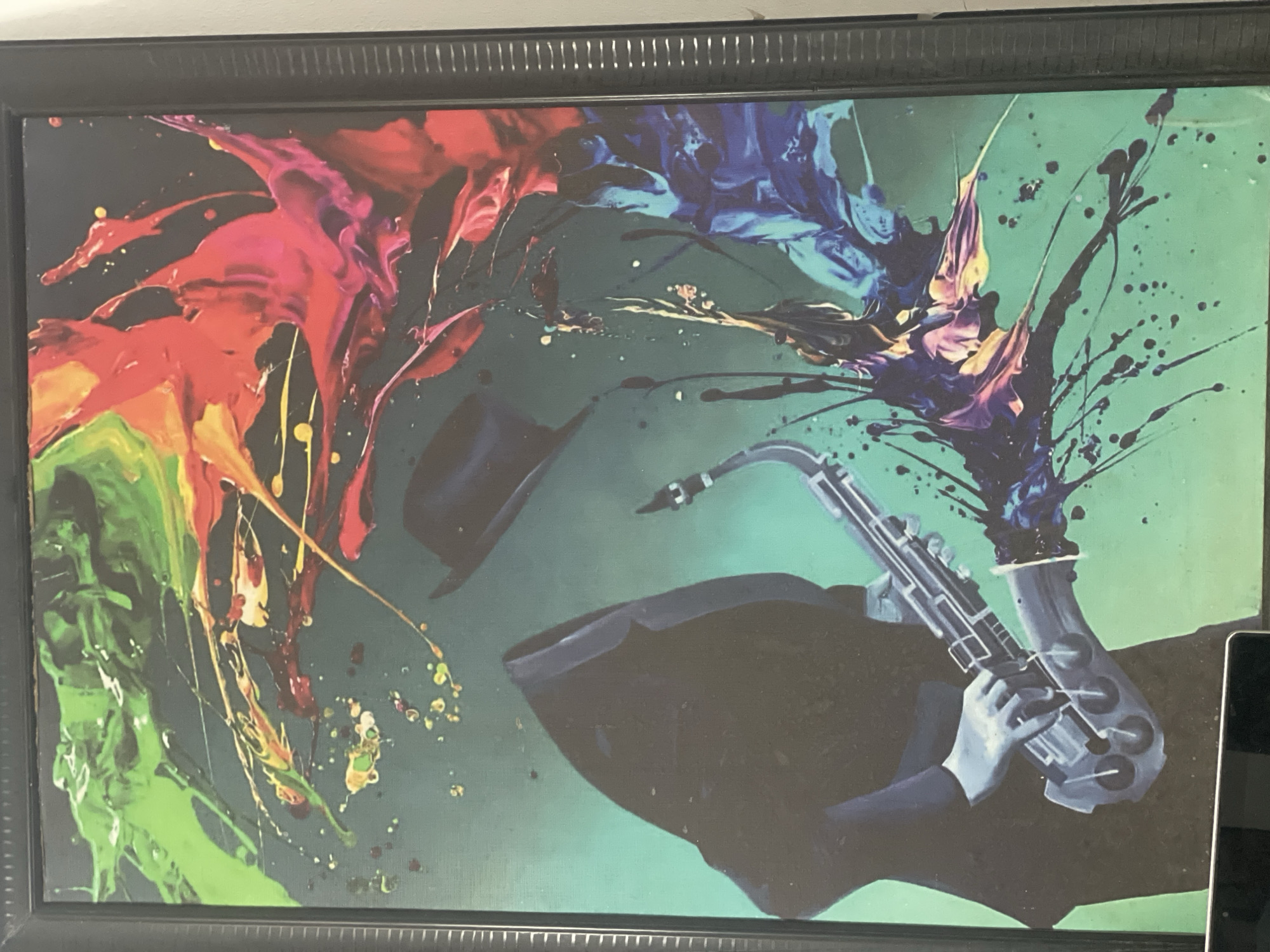 The image is a framed painting. The painting depicts a jazz musician playing a saxophone. The figure is silhouetted and wearing a hat. The saxophone and hands are rendered in shades of gray. The background is a mottled green color. Splashes of vibrant colors, including green, red, yellow, and blue, emanate from the instrument. The painting has a modern, abstract style. There is no information regarding a location in Abuja, Nigeria in the image.