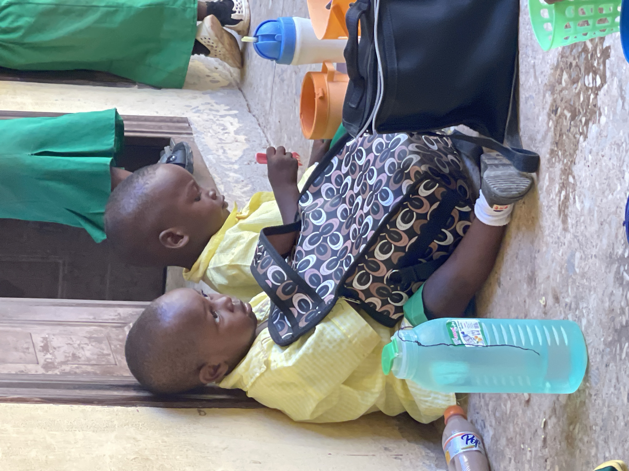 Two young children, appearing to be toddlers, are sitting on the ground in what appears to be an outdoor schoolyard or communal area in Akwanga, Nigeria. Both children are wearing yellow collared shirts, and one is also wearing green shorts. They are seated next to each other, leaning against a weathered, pale yellow wall. In front of them are various items, including a patterned bag, a large blue water bottle, an orange container, and a white and blue water bottle. The ground beneath them is paved with a speckled, light-colored material, showing some dark staining, possibly from water or dirt. In the background, other children or adults are visible, with two individuals wearing bright green skirts, suggesting a school uniform. The overall impression is one of a break time or a gathering for young students.