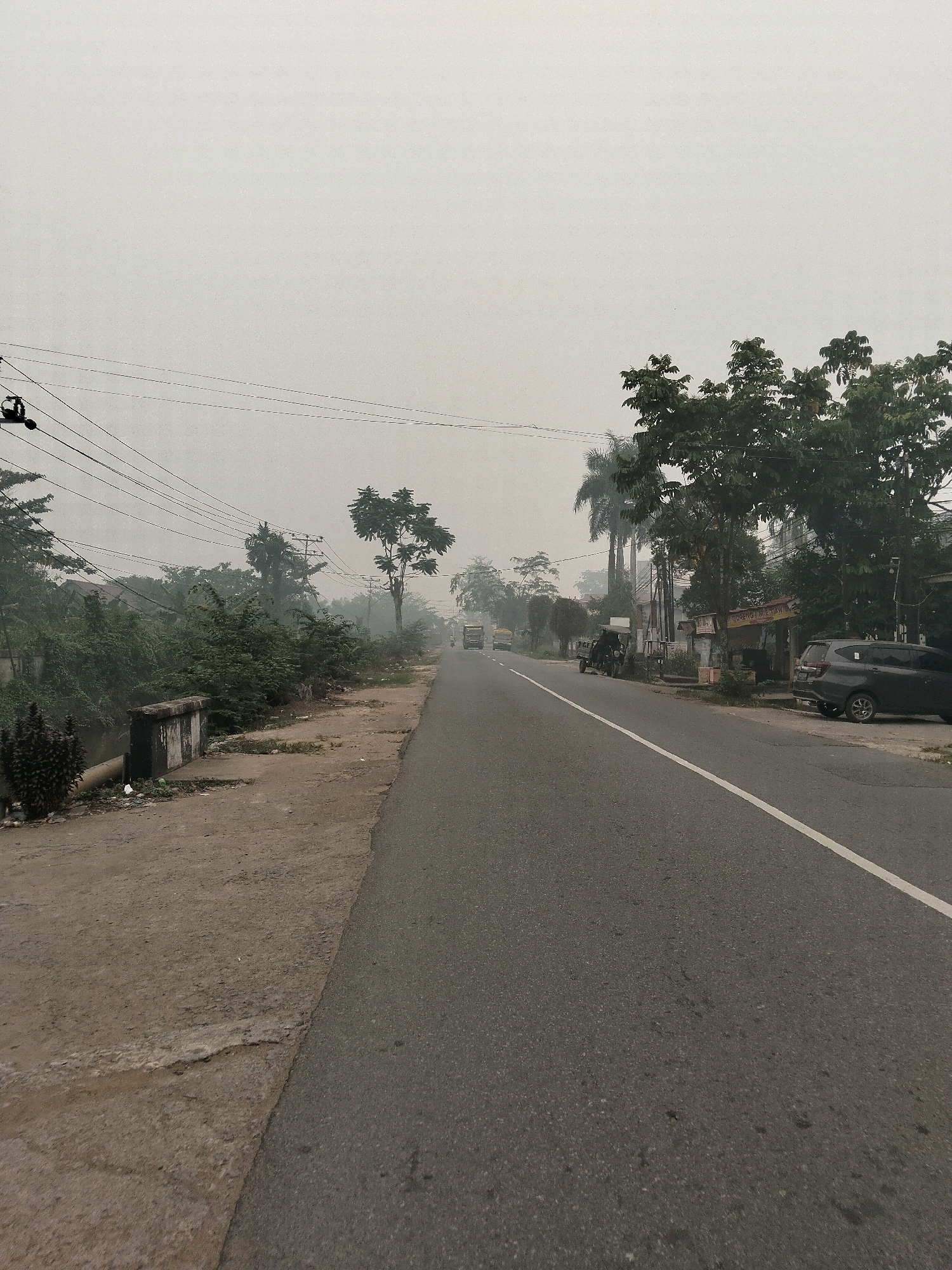 The image depicts a street scene in Teluk Kapuas, Indonesia, characterized by overcast skies and a hazy atmosphere. A paved road with a white lane divider runs through the center of the frame, curving slightly to the right. The road is flanked by a dirt and concrete walkway on the left and a line of trees and buildings on the right.
On the left side of the road, there is a narrow canal or ditch bordered by a low concrete structure and dense green foliage. Overhead, a network of power lines crisscrosses the sky, supported by utility poles. In the distance, several vehicles are visible on the road, including trucks and a car. A small, open-sided structure, possibly a roadside stall, is present on the right side, with a dark-colored SUV parked in front of it. The overall impression is that of a typical, somewhat unkempt, rural or semi-urban street in Indonesia, affected by weather conditions that obscure visibility.