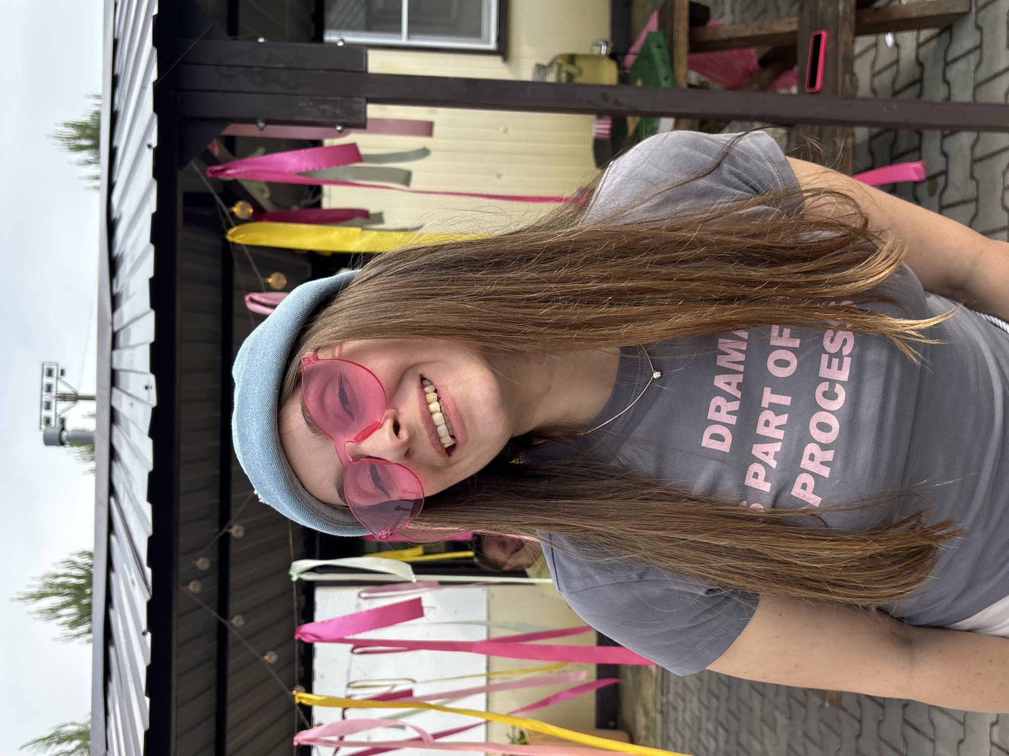 The image shows a young woman with long brown hair and pink heart-shaped sunglasses. She is wearing a light blue beanie and a gray t-shirt with pink text that reads "DRAMA IS PART OF THE PROCESS". The woman is smiling and looking at the camera.
She is outdoors, possibly at a party or outdoor gathering, with colorful ribbons and string lights in the background. Behind her, there is a building with a dark roof and light-colored siding, and some greenery is visible on the left side of the frame. The weather appears to be overcast. The paving stones on the ground suggest an outdoor patio or courtyard. The scene conveys a sense of fun and carefree emotion.