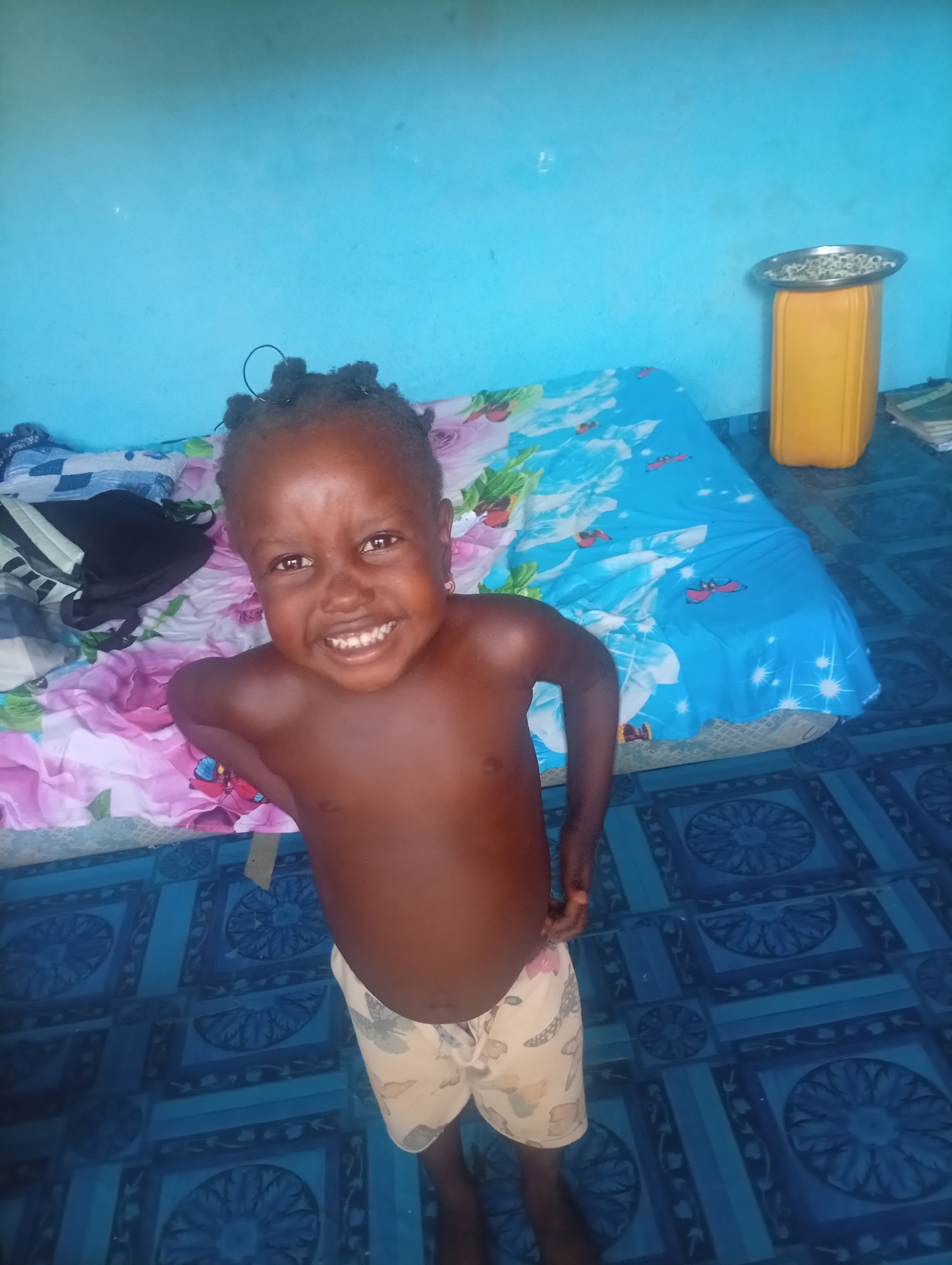 A young, dark-skinned child smiles at the camera. The child stands in a room with light blue walls and blue patterned tile flooring. A bed covered in a pink floral and blue patterned sheet is behind the child. A yellow plastic container with a metal tray on top is in the background. A black backpack sits on the bed. Several books sit on the floor behind the yellow container.