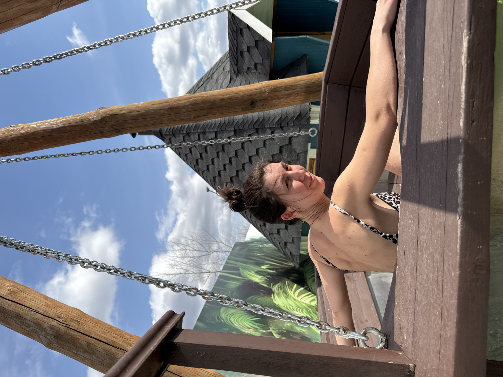 A woman with brown hair pulled into a bun is shown from the back in a leopard print bikini. She is holding onto the side of a wooden structure. The background reveals a structure with a grey shingled roof, a blue accent, and a green backdrop featuring a plant design. Chains are visible, seemingly part of the structure the woman is holding. The location is Votkinsk, Russia.