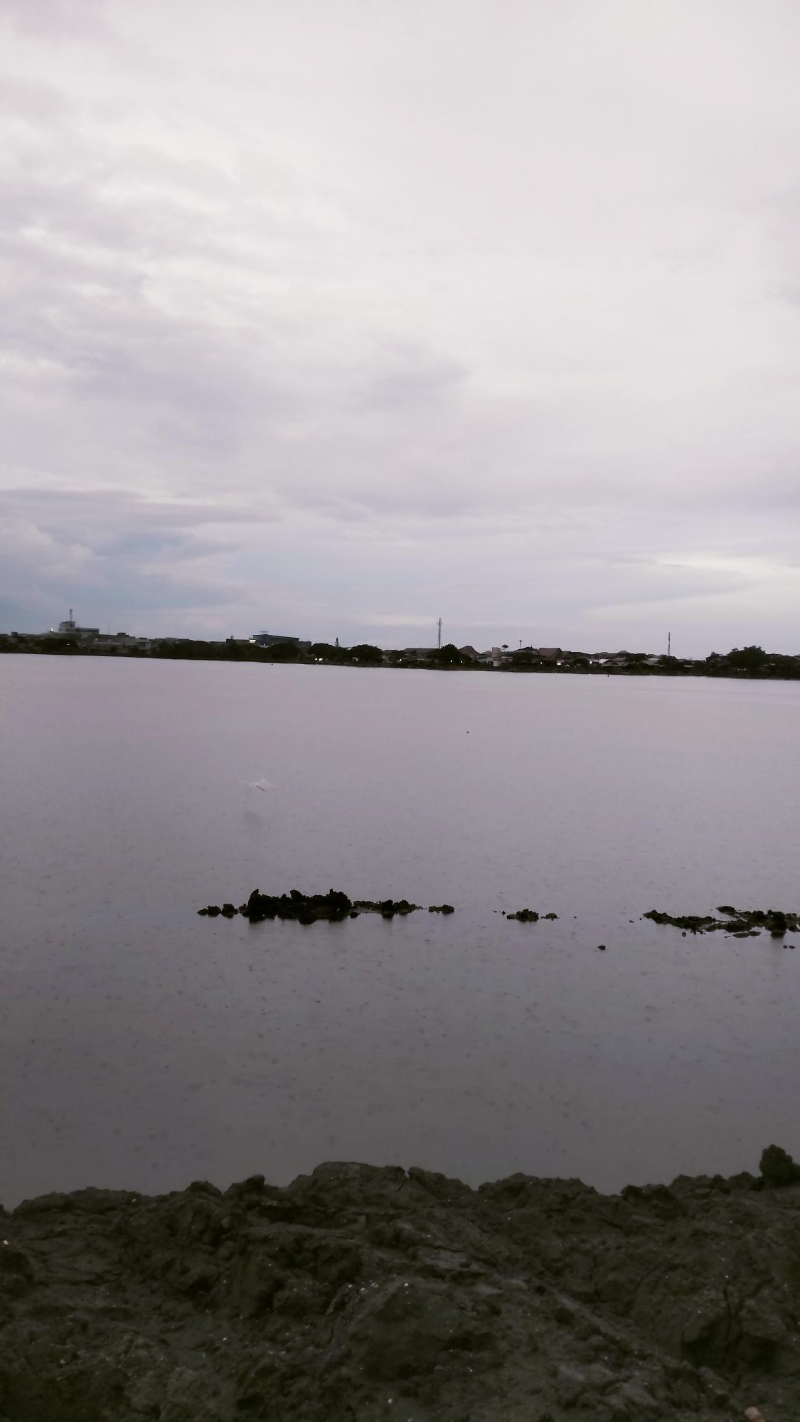 The image captures an overcast scene of a body of water in Lhokseumawe, Indonesia, likely a lake or estuary. The sky is dominated by thick, light gray clouds, indicating a cloudy or potentially rainy day.
The central feature is a wide expanse of calm water, its surface speckled with visible ripples or indentations, strongly suggesting light rain is falling. A short, irregular line of dark rocks or debris emerges from the water in the mid-ground, breaking the otherwise smooth surface.
Along the distant horizon, a subtle skyline of buildings and trees is visible, indicating a populated area across the water. The structures appear low-rise, with various roofs and outlines, some blending into the distant tree line. One building on the far left seems slightly more prominent than the others.
In the immediate foreground, a dark, rugged bank of earth or mud occupies the bottom portion of the frame. This bank is uneven and textured, contributing to the natural, unrefined feel of the landscape.
The overall impression is one of quiet serenity, with the muted colors and cloudy conditions creating a somewhat somber or contemplative atmosphere. The absence of direct sunlight suggests it could be early morning, late afternoon, or simply a heavily overcast day. No visible text is present in the image.