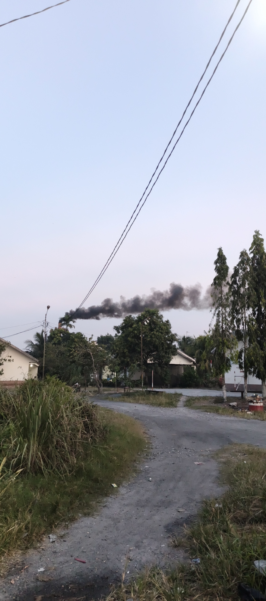 This image depicts a rural scene in Danau Lamo, Indonesia, characterized by a dirt road winding through a sparsely populated area. In the background, a plume of dark smoke billows into the sky from what appears to be a chimney or stack, suggesting industrial or agricultural activity. Several modest houses and trees are visible, interspersed with tall grasses and vegetation. Overhead, power lines stretch across the frame, indicating the presence of electricity infrastructure. The overall impression is one of a quiet, developing community with hints of industrial presence.