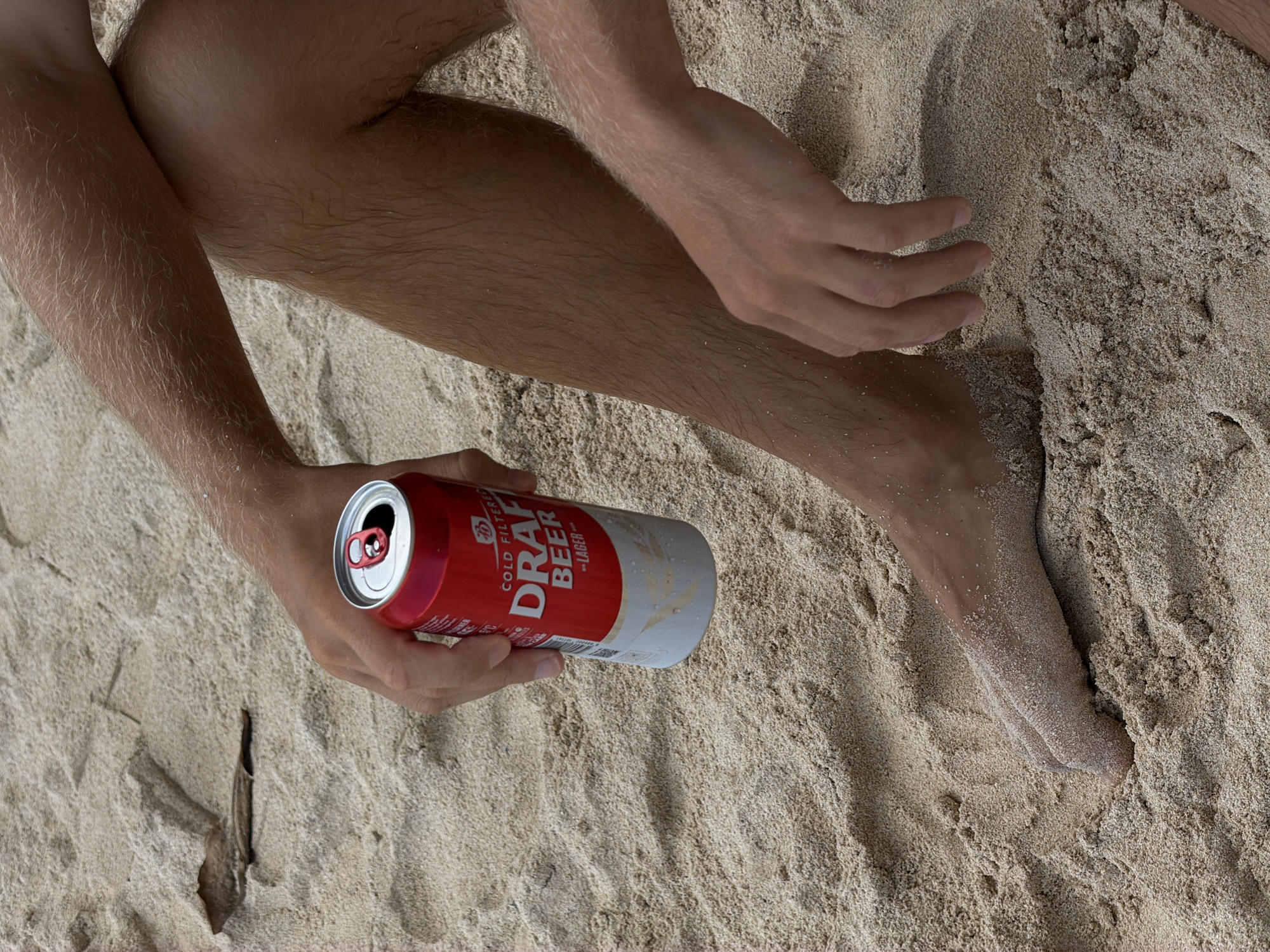 A person with fair skin and noticeable body hair is sitting on a sandy surface. The person's legs and arms are visible, positioned near a can of "Cold Filtered Draft Beer - Lager" in red and white packaging. The can appears to be open and partially consumed. The sand is light in color with varying textures, suggesting a natural outdoor setting. The location is in Kuta Selatan, Indonesia, based on the beach setting of the image.
