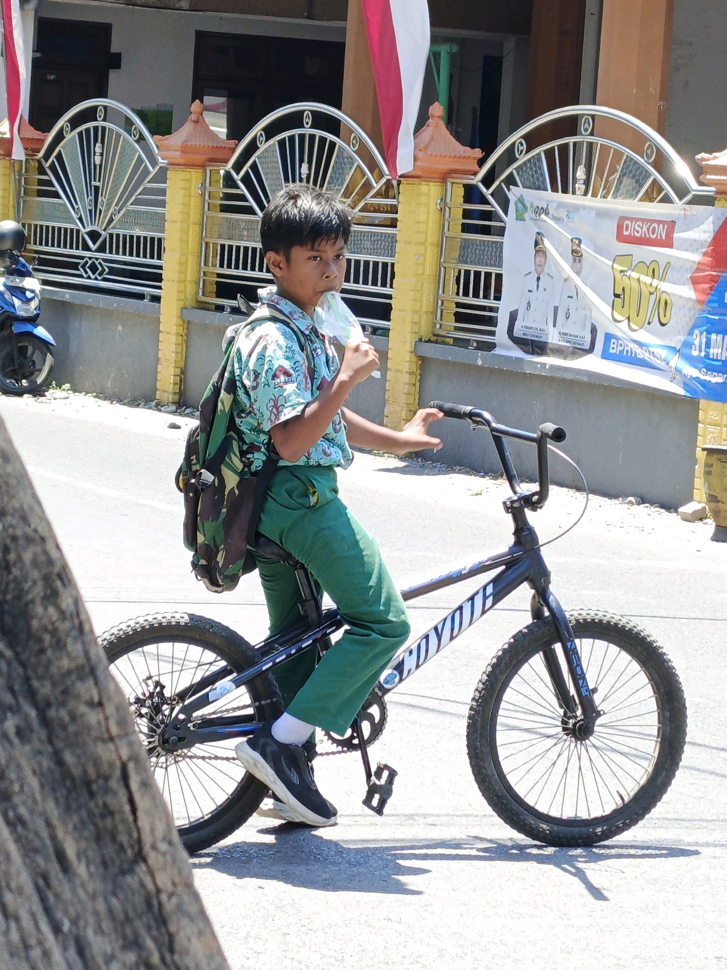 A young boy, dressed in a light blue, patterned short-sleeved shirt and green trousers, is riding a black BMX bicycle on a paved street in Ketegan, Indonesia. He has a camouflage backpack on his back and is holding a white object, possibly a face mask, to his mouth. The bicycle has "COYOTE" written in white on its frame. In the background, there is a yellow-and-orange patterned fence with decorative elements and a building behind it. A blue motorcycle is parked to the left of the frame, and a promotional banner with text including "DISKON" and "50%" is visible on the fence. The sun is shining, casting shadows on the ground. The overall scene depicts a street view with a boy engaged in cycling.