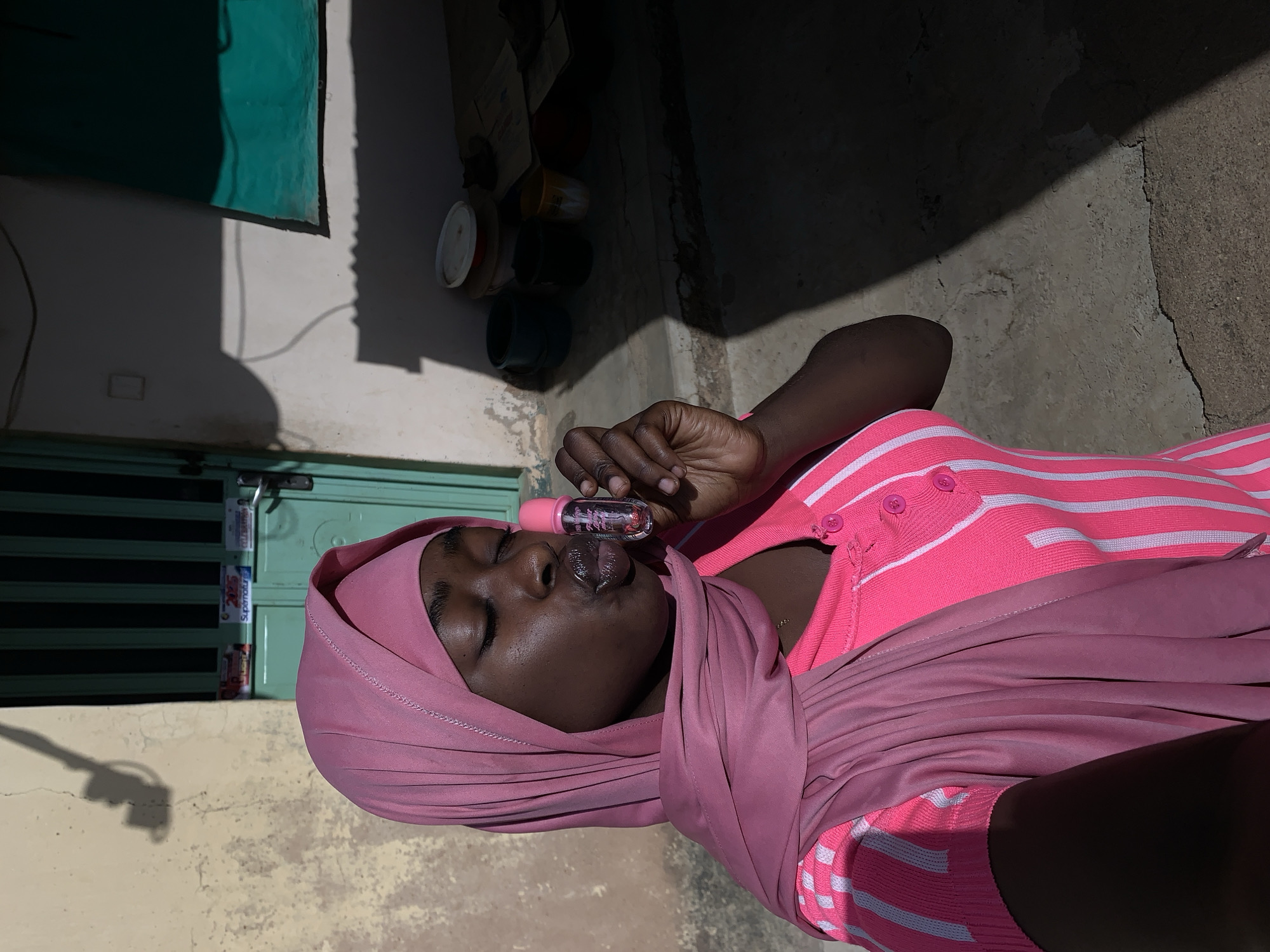 A woman with dark skin is lying on the ground in Rafin Par, Nigeria, wearing a pink headscarf and a pink and white striped shirt. She is holding a small, clear bottle of liquid to her lips. The background includes a concrete wall, a green door, and various objects on a wall. The lighting is bright and creates strong shadows.