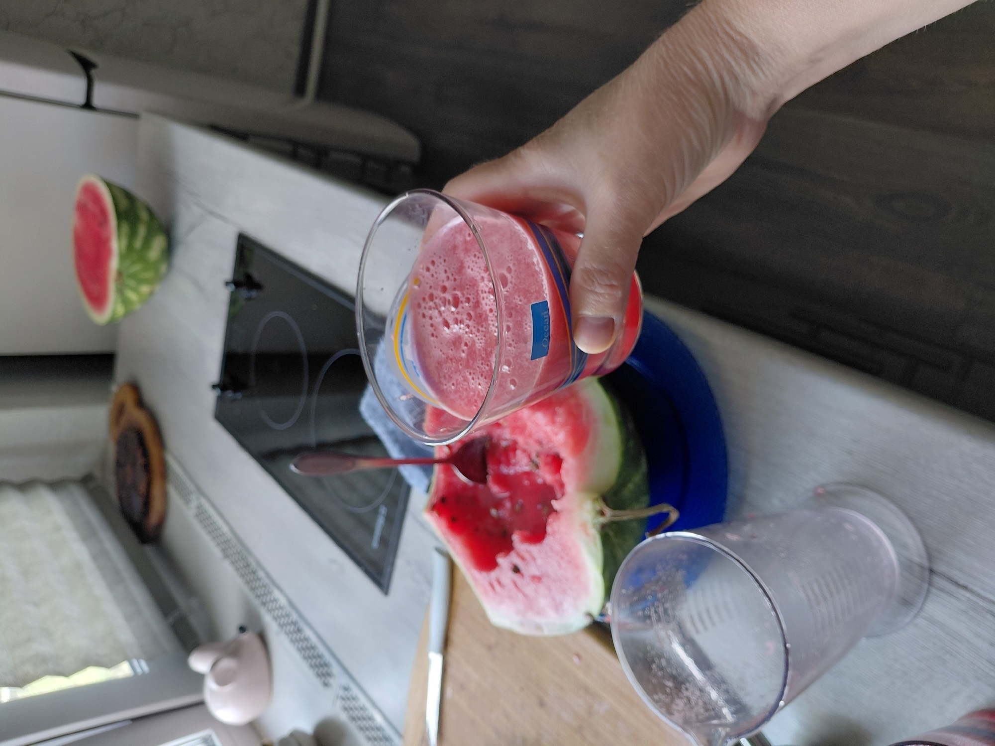 The image depicts a domestic kitchen scene, possibly in Novosibirsk, Russia. A person's hand is holding a glass of pink juice, which appears to be watermelon juice, to the viewer. In the foreground, a half-eaten watermelon sits on a blue bowl, with a spoon in it. To the left, a measuring cup is visible. In the background, a stove top and kitchen counter are visible, with a small decorative pink object resembling a mouse. The overall impression is of a casual, home-based food preparation activity.