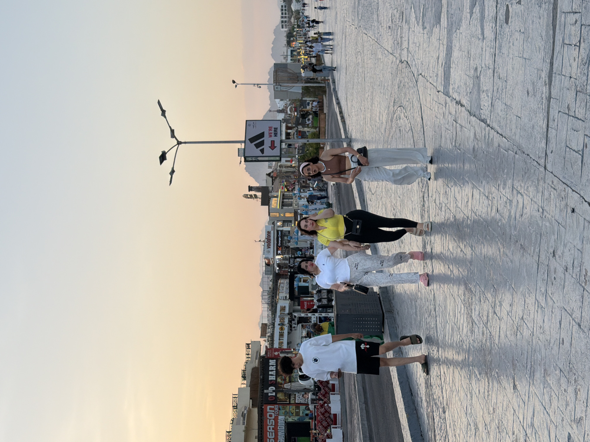 In Sharm El Sheikh, Egypt, at dusk, four individuals walk along a paved promenade. From left to right: a young man wearing a white t-shirt, black shorts, and sandals, looking at his phone; a woman in a white polo shirt and patterned pants with pink slides, carrying a black purse; another woman in a yellow t-shirt and black leggings with white slides, also carrying a black purse; and a fourth woman in a brown crop top, white wide-leg pants, and white sneakers, wearing a headband and carrying a black purse. In the background, there is a street with shops and restaurants, a street lamp with a sign that reads "WE'RE HERE" with an Adidas logo and an arrow pointing left, and mountains in the distance. The sky exhibits a gradient of sunset colors, transitioning from orange near the horizon to a pale blue at the top.