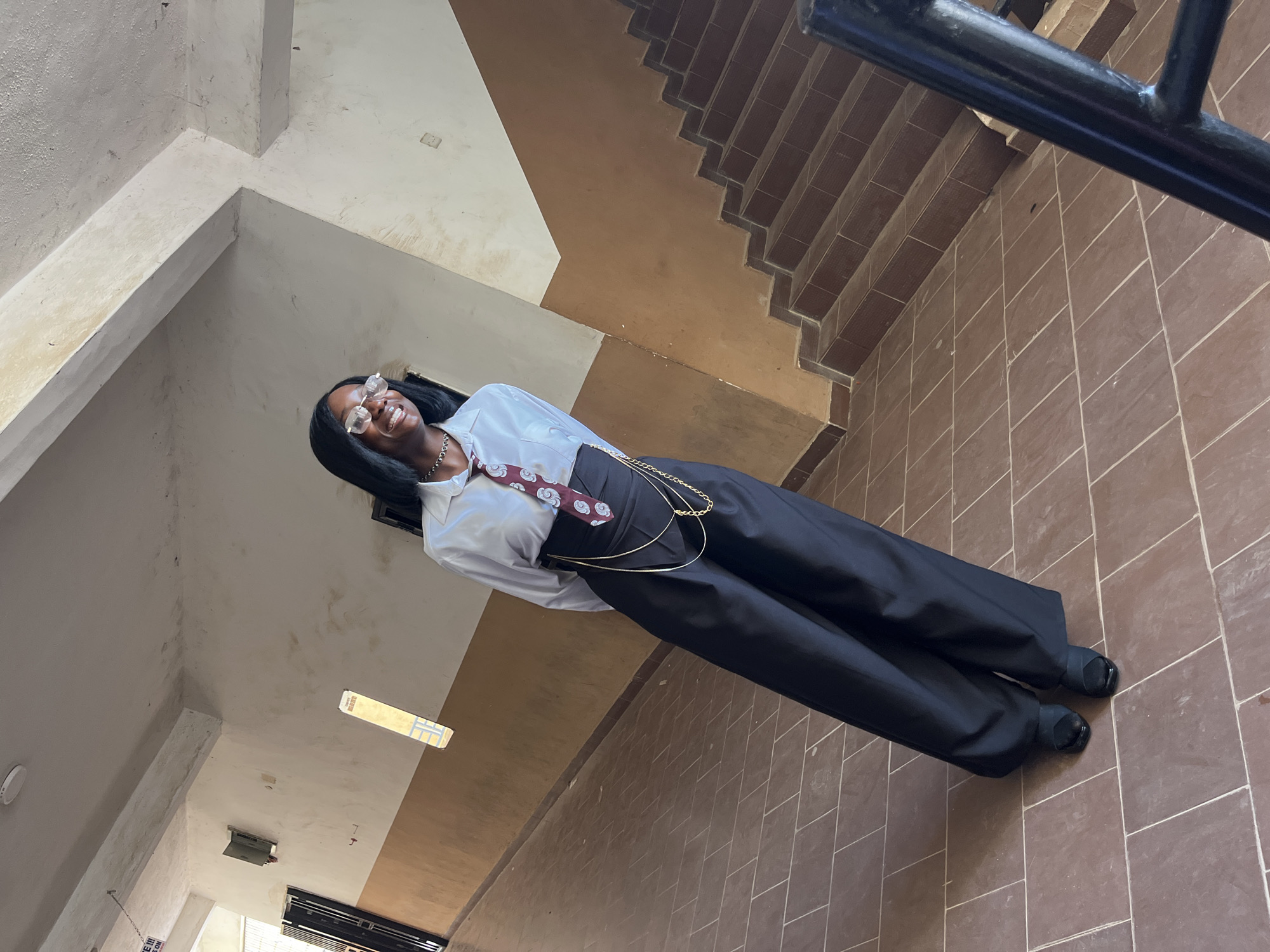 A young Black woman stands in a hallway, her body angled to her left. She wears a white collared shirt, a patterned tie, and high-waisted black pants. A gold chain is draped around her waist. She has on glasses and a necklace. The hallway has tiled floors and brown walls, with a staircase visible on the right. The scene is likely located in Jos, Nigeria, given the architectural style and the presence of a young Black woman.