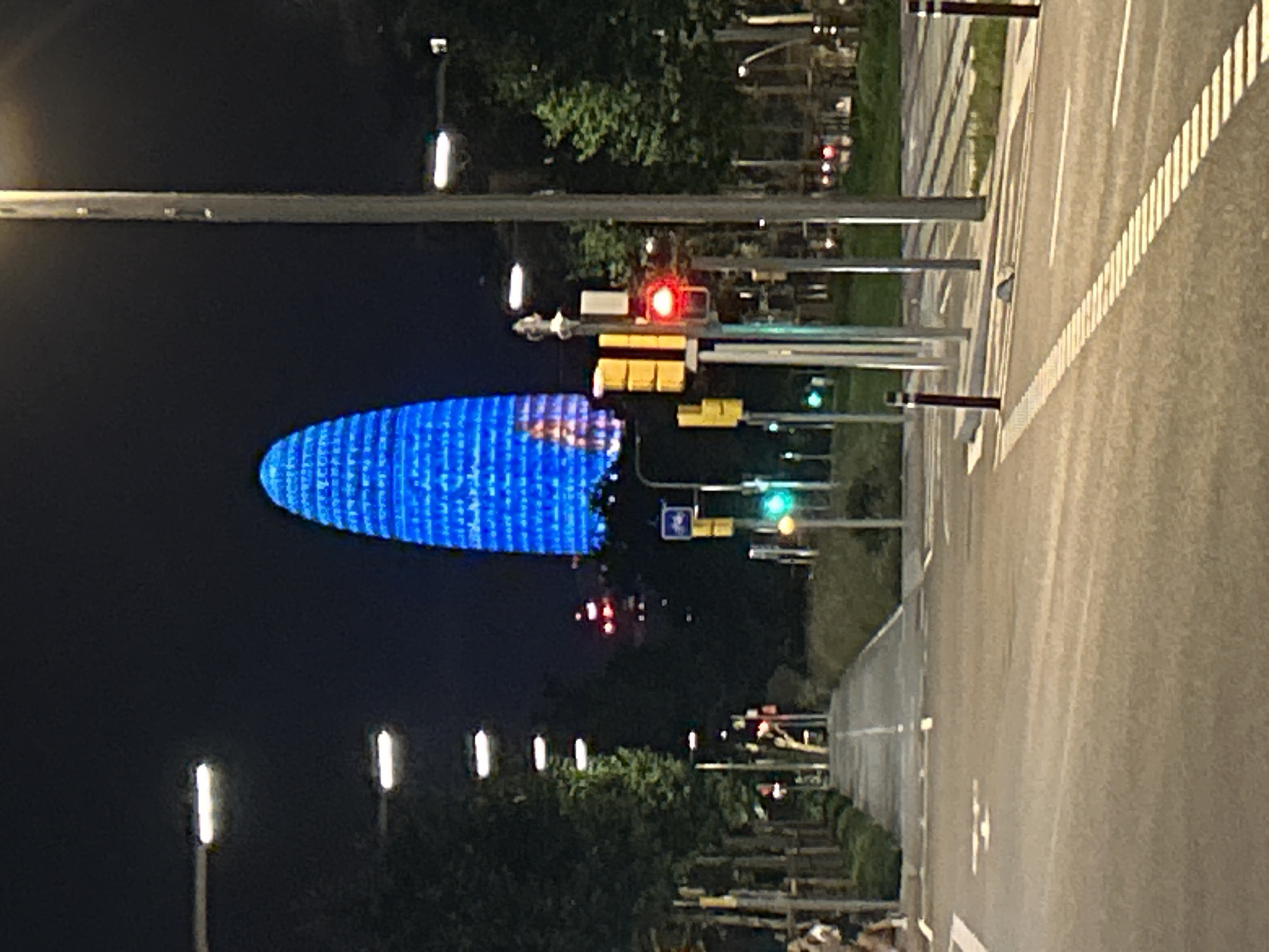 The image depicts a night scene in Barcelona, Spain, featuring the iconic Torre Glòries skyscraper illuminated in vibrant blue. The skyscraper is the dominant subject in the background, appearing as a luminous, oval-shaped structure against the dark night sky. In the foreground, a street or road is visible, with marked lanes and pedestrian walkways. Traffic lights are present, with one prominently displaying a red light. Streetlights line the sides of the road, casting light onto the surroundings. Trees and greenery are interspersed along the street, adding to the urban landscape. A few individuals are partially visible on the left side of the image, walking or standing. The overall ambiance is urban and modern, characteristic of a city at night.