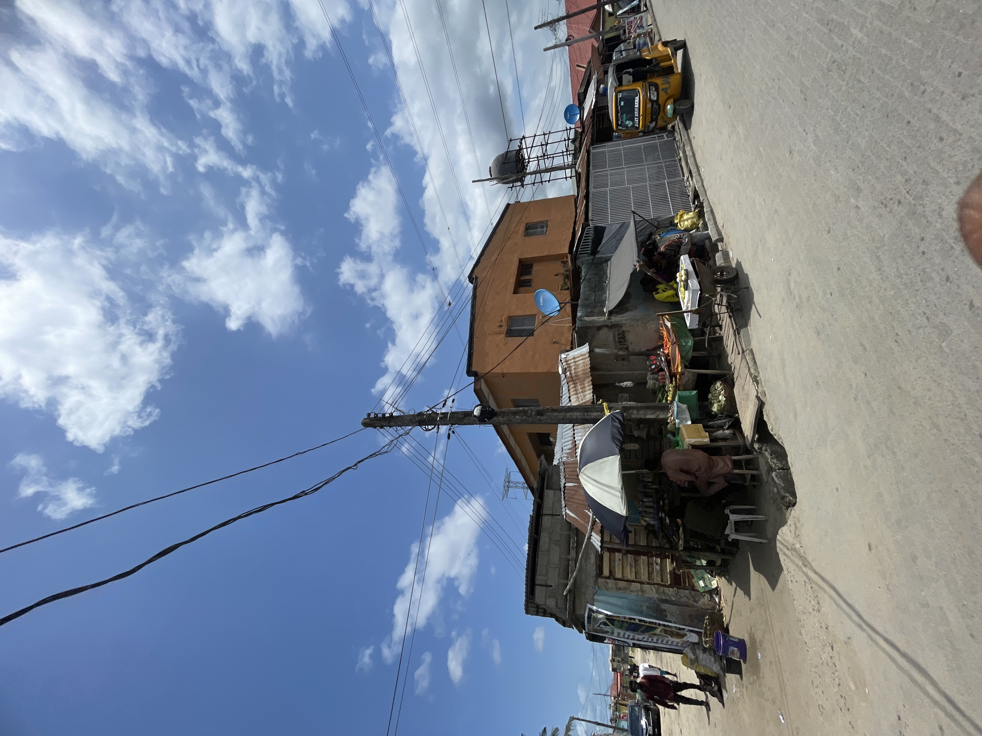 The image depicts a street scene in Ado, Nigeria, under a bright sky with scattered clouds. A two-story, orange building with a water tank and satellite dishes stands adjacent to the road. Utility poles with power lines stretch across the sky.
Along the street, there are informal vendors selling produce and other goods from makeshift stalls covered with umbrellas. A yellow auto-rickshaw (keke) is parked near one of the stalls. Pedestrians are walking along the street. The road surface is cracked and uneven.