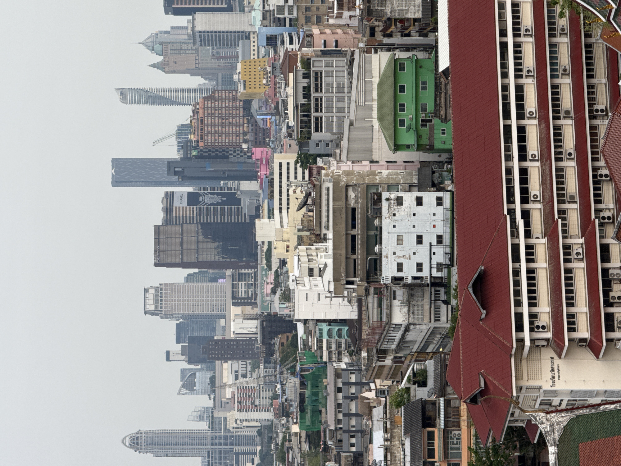 The image depicts an aerial view of Bangkok, Thailand, characterized by a dense urban landscape. Skyscrapers dominate the left side of the frame, gradually giving way to mid-rise buildings and low-rise residential structures towards the right. The overall environment suggests a hazy atmosphere, typical of urban areas with high levels of air pollution.
Notable landmarks include the distinctive shapes of several skyscrapers, such as the Mahanakhon, a modern tower with a pixelated facade, and other high-rise buildings with varying architectural styles. Interspersed among the modern structures are older, traditional Thai buildings with tiled roofs and characteristic architectural details.
The composition highlights the juxtaposition of modern high-rises and older, smaller structures, showcasing the diverse architectural styles of Bangkok. There are no visible people or activities depicted in the scene.