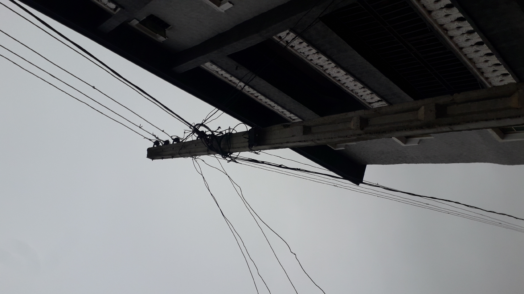 The image shows a view of an outdoor scene, likely in an urban area. A concrete building overhangs the shot, with a corrugated metal roof or awning extending from it. Attached to the building and extending into the sky is a concrete utility pole that is laden with numerous wires. The wires are connected in bundles and are strung across the grey sky. The weather appears overcast or foggy. The location of the image is likely the city of Nkpor, Nigeria.