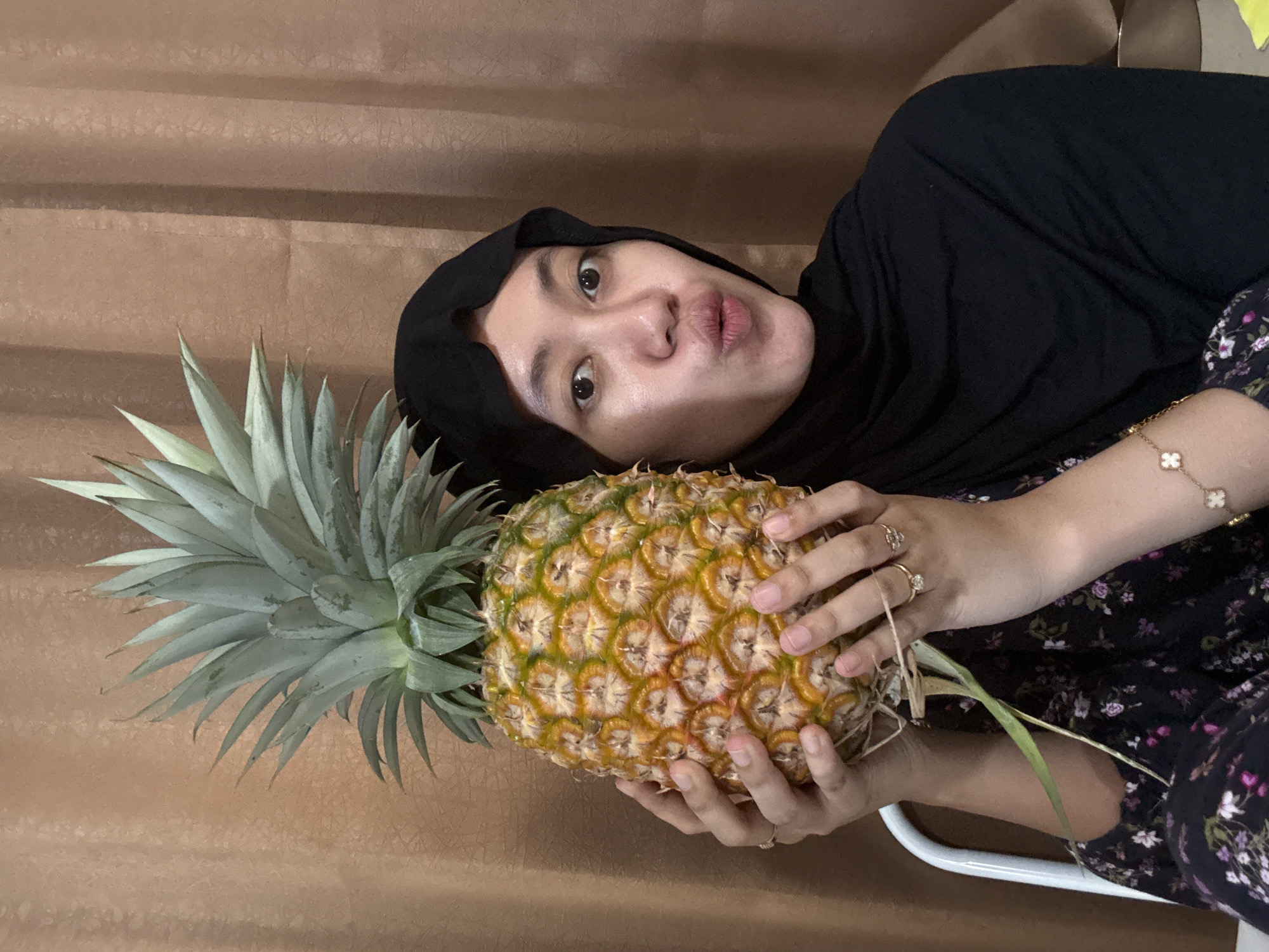 A person wearing a black hijab and a floral patterned top is holding a pineapple in both hands. The person is puckering their lips and looking at the camera. They are wearing rings on their fingers and a bracelet on their wrist. The background is a brown curtain. The location is Gadingrejo, Indonesia.