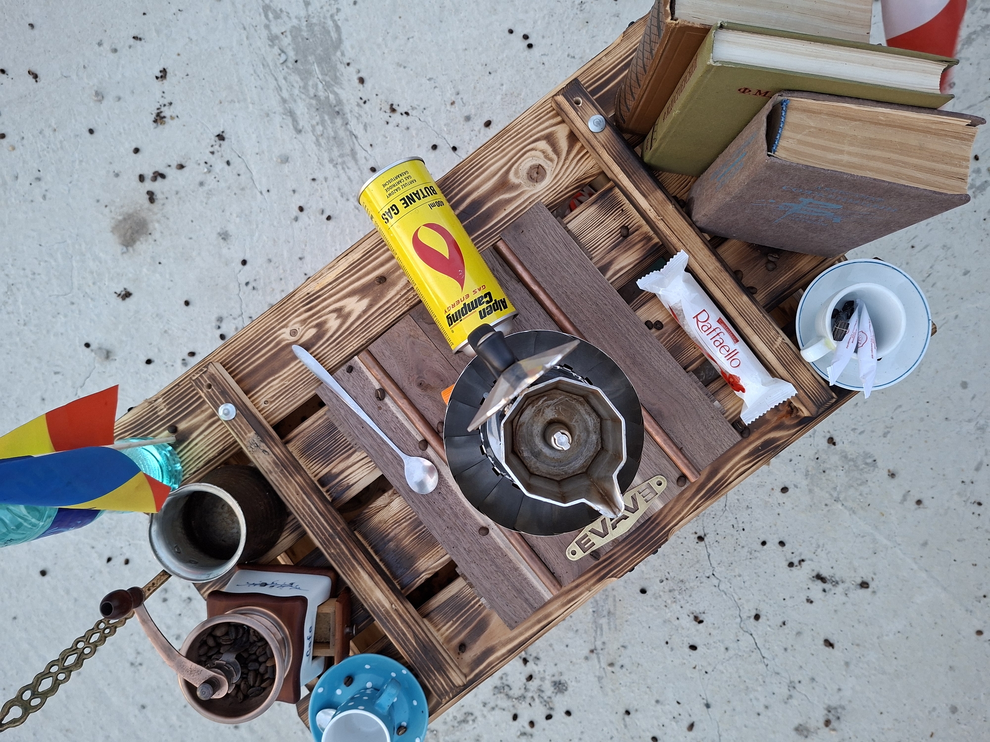 The overhead shot depicts a rustic wooden pallet serving as a table on a concrete surface, likely in the city of Sociteni, Moldova. Scattered coffee beans litter the ground and the pallet. On the pallet rests a vintage copper coffee grinder filled with beans, a small metal pot, a blue and white polka-dotted teacup and saucer, a metal spoon, and a Moka pot. To the right, a yellow butane gas canister is visible, along with a wrapper for a Raffaello candy. Stacked against the side of the pallet are several old books. Two small Romanian flags, attached to a blue bottle, stand upright. The overall scene suggests an outdoor coffee preparation setting with a vintage aesthetic.