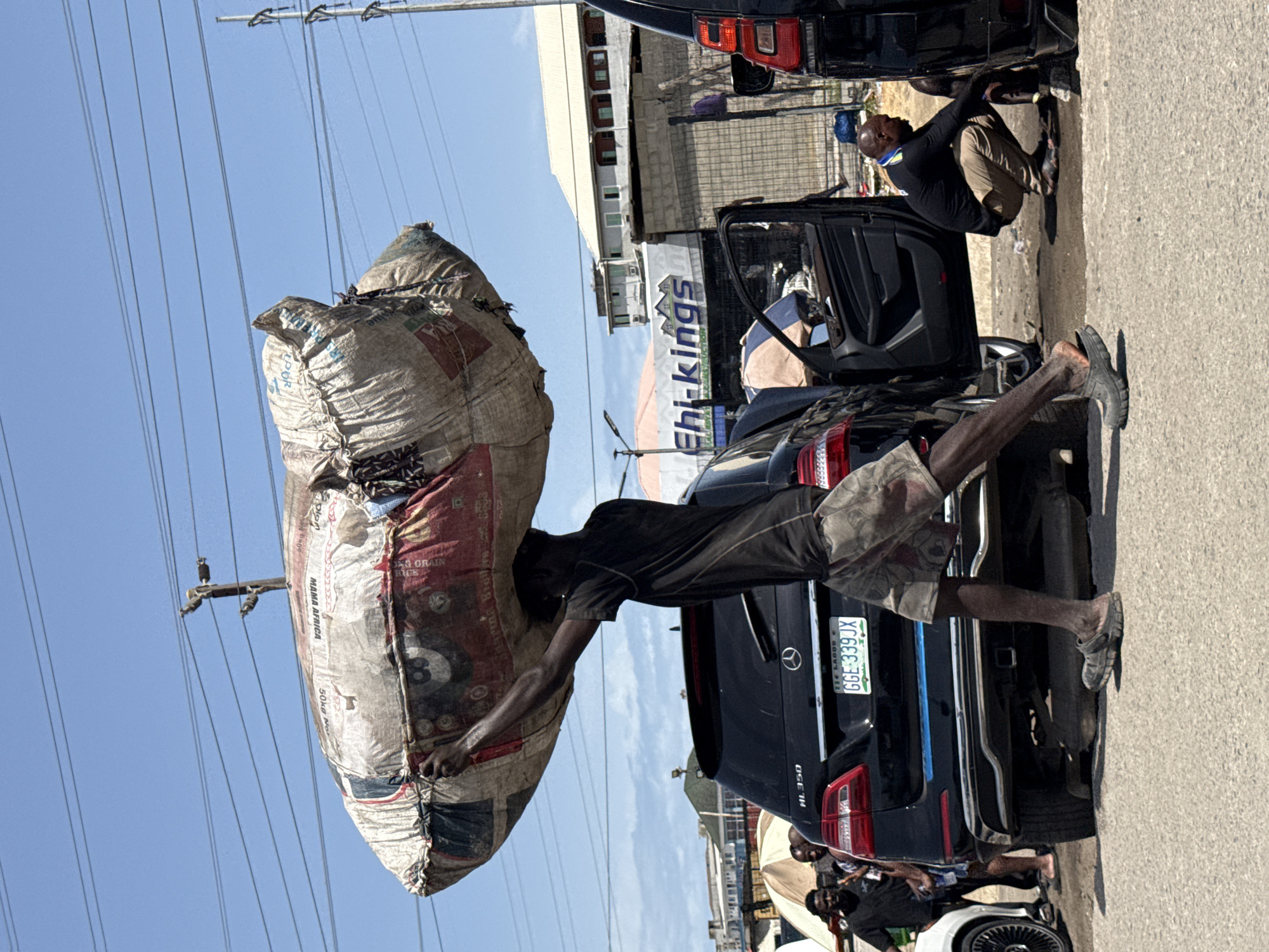 A man in casual clothing is seen carrying a large sack on his shoulder while standing on the back of a black Mercedes-Benz ML 350 SUV with license plate GGE-339JX registered in Lagos, Nigeria. Another man is kneeling near the open car door. The backdrop includes power lines, a building, and a sign reading "Ehi-kings." The location is Igboefon, Nigeria.