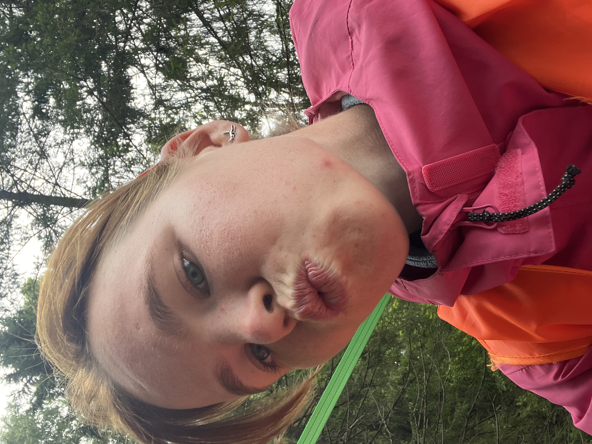 This is a selfie of a young person wearing a pink and orange jacket outdoors. The subject is making a kissing face, with their lips pursed. They have light-colored hair and a small silver earring in their left ear. The background shows green trees and a cloudy sky, suggesting it is daytime. The person is wearing an orange life vest under their jacket. The image is oriented sideways. The location context provided is Сергино, Russia.