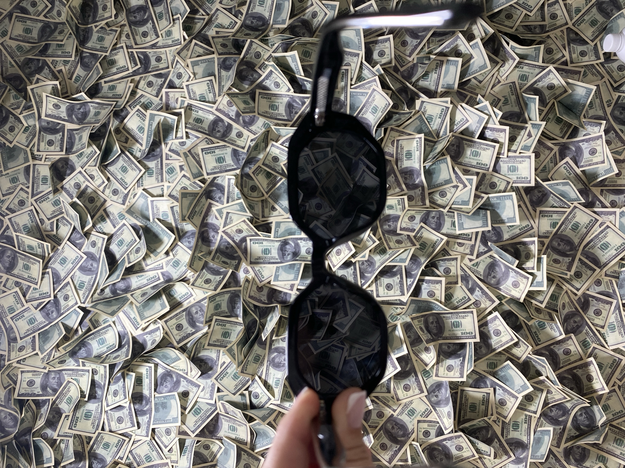 A high-angle shot depicts a dense spread of United States one hundred dollar bills, obscuring most of the surface beneath. A hand holds a pair of dark sunglasses above the money, reflecting the bills within the lenses. The background is filled with more of the same currency. The location, Sochi, Russia is not discernible from the visual content of the image.