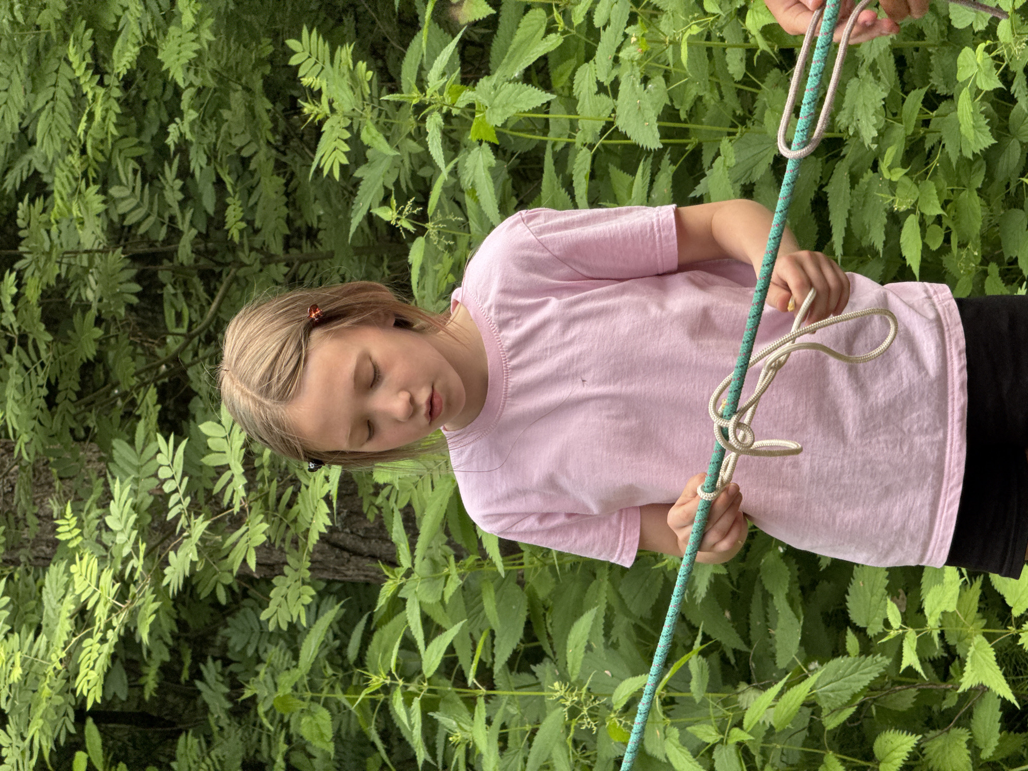 A young girl with blonde hair, wearing a pink t-shirt and black shorts, stands outdoors in a lush green environment with many plants. She is focused on tying a knot with two ropes. One rope is thick and green with flecks of red and white, and she is holding it horizontally. The other rope is thinner and off-white, and she is in the process of knotting it around the green rope. Another person's hands are visible on the right side of the image, also holding the green rope, suggesting they are participating in the activity with the girl. The background is filled with dense foliage, indicating a natural or park-like setting. No specific identifiable landmarks of Chaikovsky, Russia, are visible in the image.