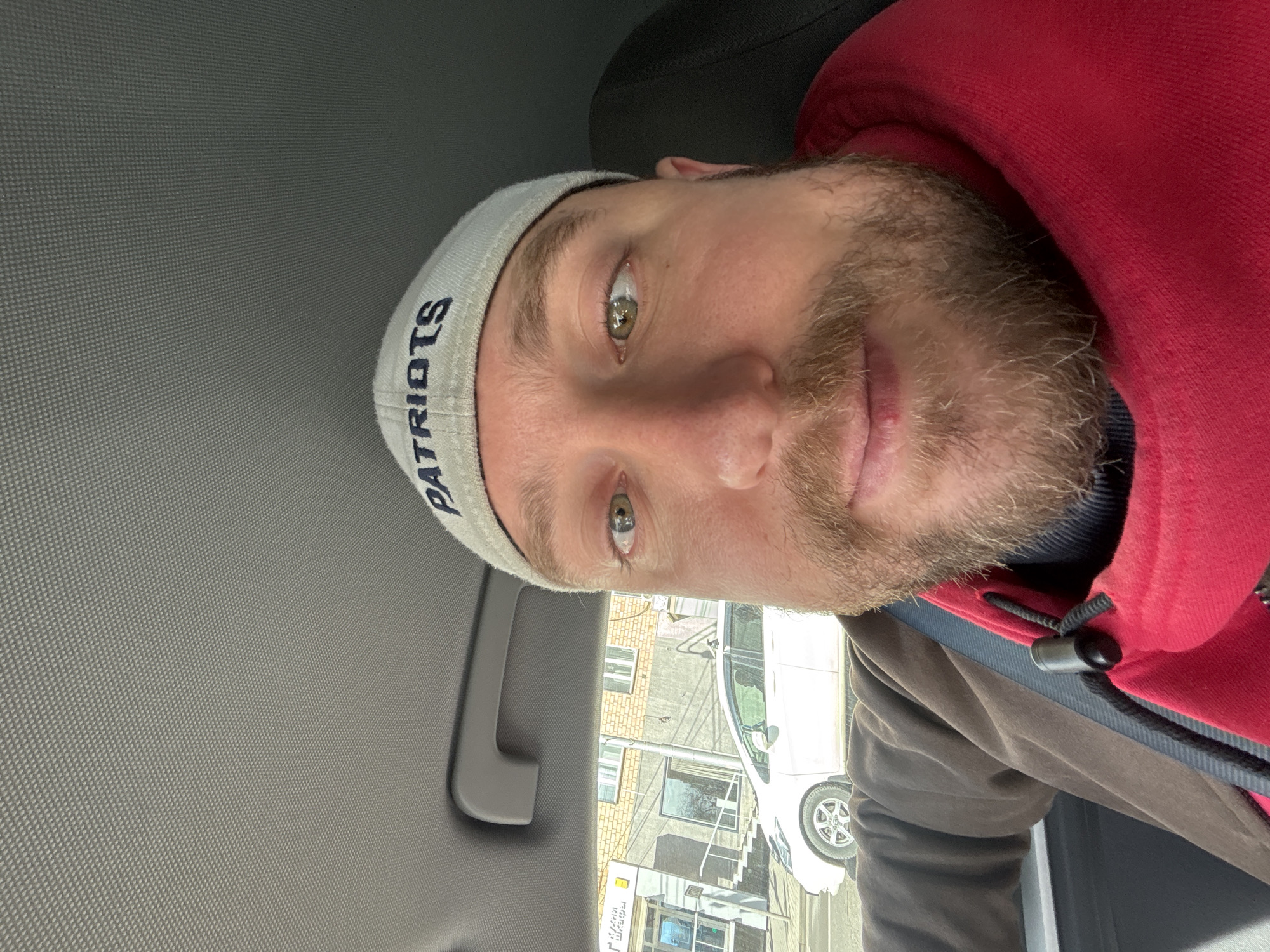 A man with light skin, a beard, and hazel/green eyes is captured in a selfie taken inside a car. The image is oriented sideways, with the man's head tilted to the left. He is looking directly at the camera with a neutral expression. He wears a light grey beanie with "PATRIOTS" written in dark blue letters, a red hooded sweatshirt, and a brown jacket over a dark blue underlayer.
The setting appears to be the interior of a vehicle, with a grey fabric ceiling/headliner and a grey grab handle visible above and to the left of the man. A dark headrest is behind him. Through the car window, an outdoor scene is visible. A white car is parked or driving on a street, with parts of brick and lighter-colored buildings in the background. A metal pipe runs vertically down one of the buildings.
Visible text includes "PATRIOTS" on the man's beanie and, through the car window, a partially obscured and possibly mirrored sign with Cyrillic text that appears to read "Лайм Шакья" (Lime Shakya). The presence of Cyrillic script and the provided context indicate the location is Krasnoyarsk, Russia. The lighting suggests it is daytime with natural light.